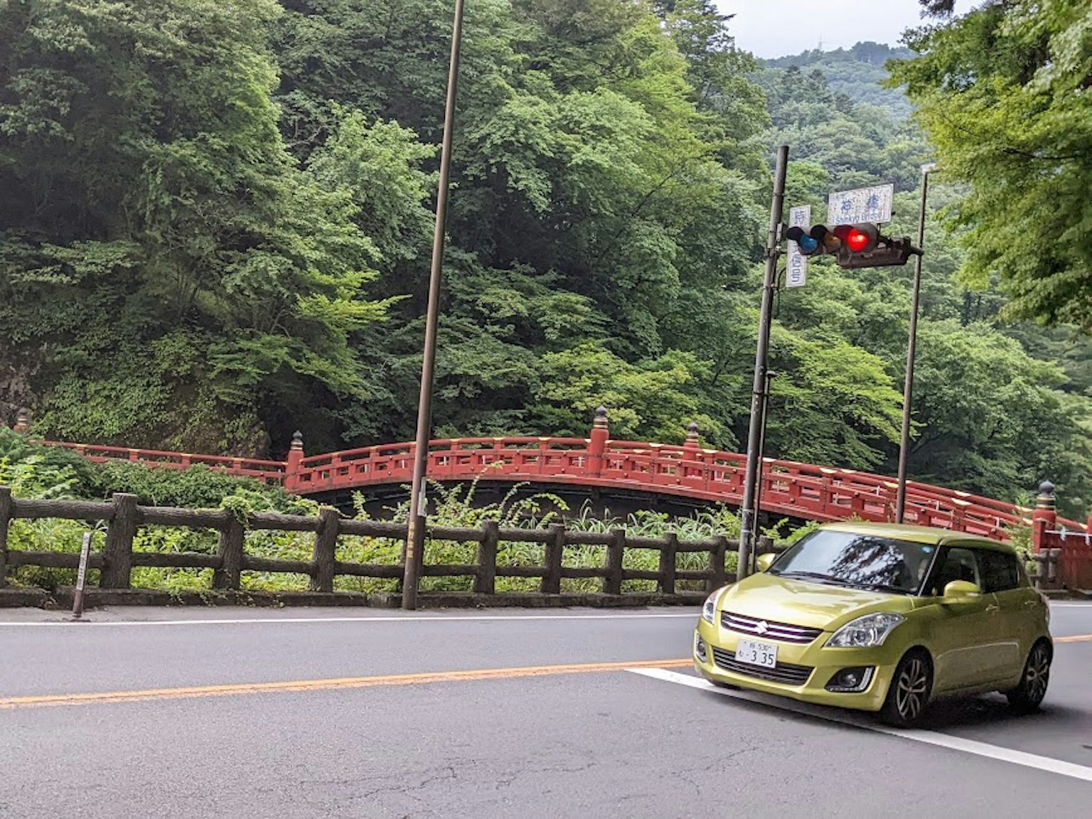 A green car passing in front of a red bridge surrounded by lush greenery