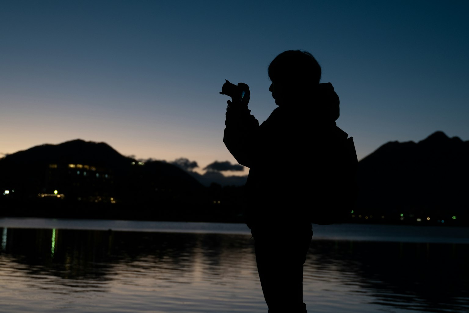 Silhouette einer Person mit einer Kamera am Ufer des Sees bei Dämmerung