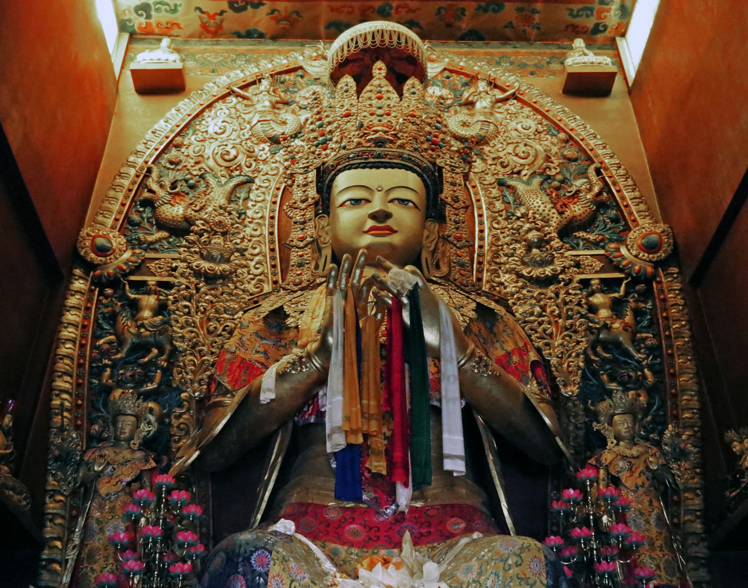 รูปปั้นพระพุทธรูปสีทองนั่งอยู่บนแท่นบูชาพร้อมมือที่ทำท่ากราบไหว้ล้อมรอบด้วยริบบิ้นหลากสีและดอกไม้