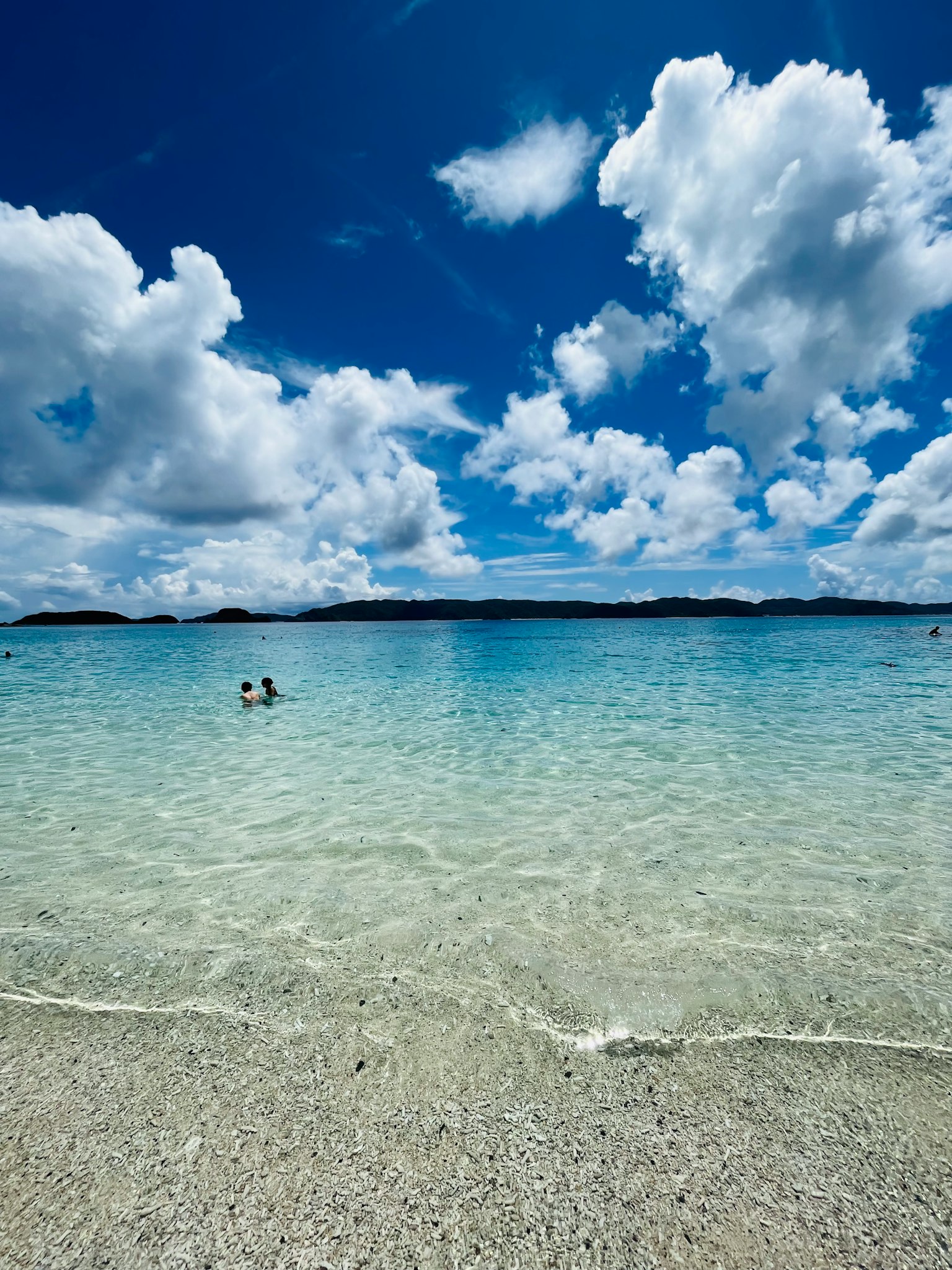 Clear turquoise water under a bright blue sky with fluffy white clouds two people wading
