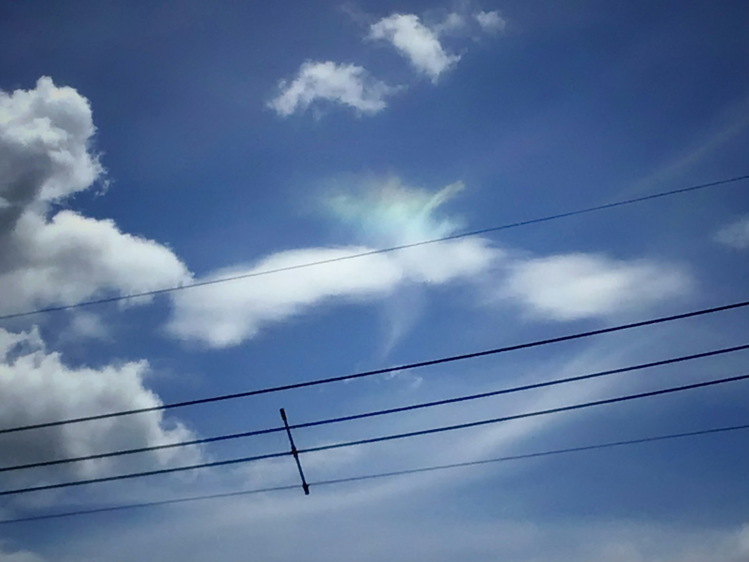 Weiße Wolken vor einem blauen Himmel mit einem regenbogenähnlichen Lichtphänomen