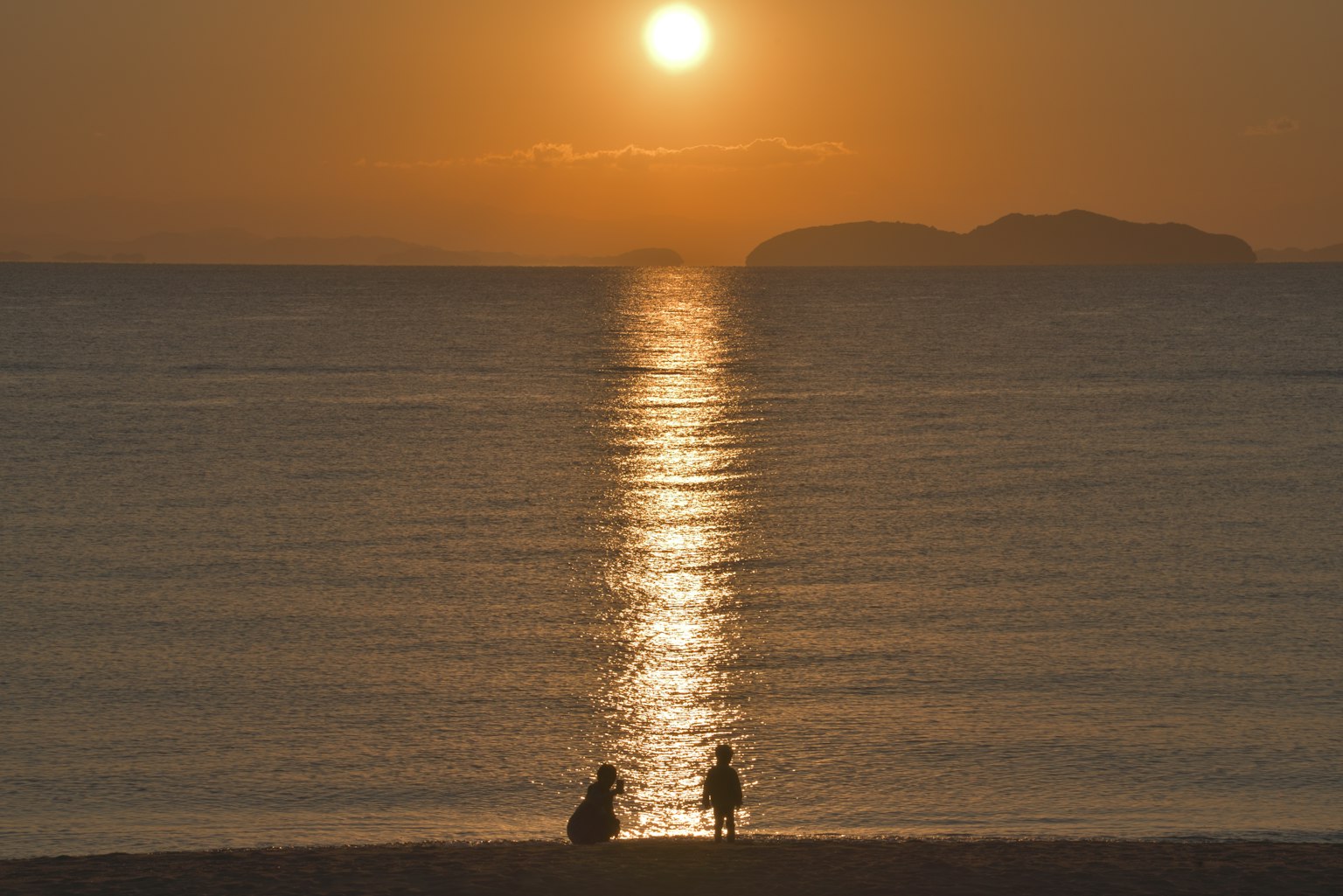 Silhouette von zwei Personen, die am Strand sitzen, mit dem Sonnenuntergang, der sich im Ozean spiegelt