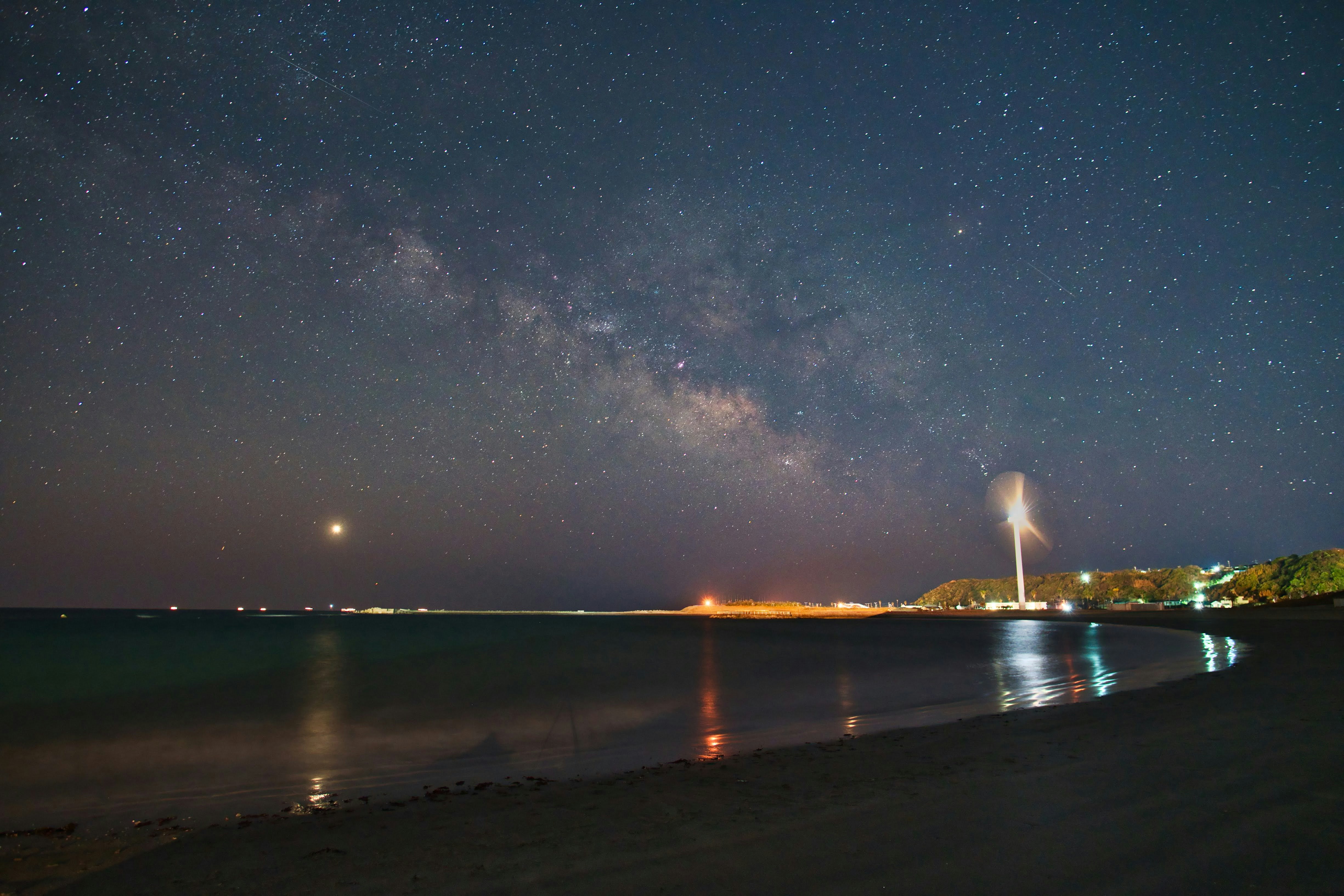 夜の海辺で光る灯台と星空が広がる風景
