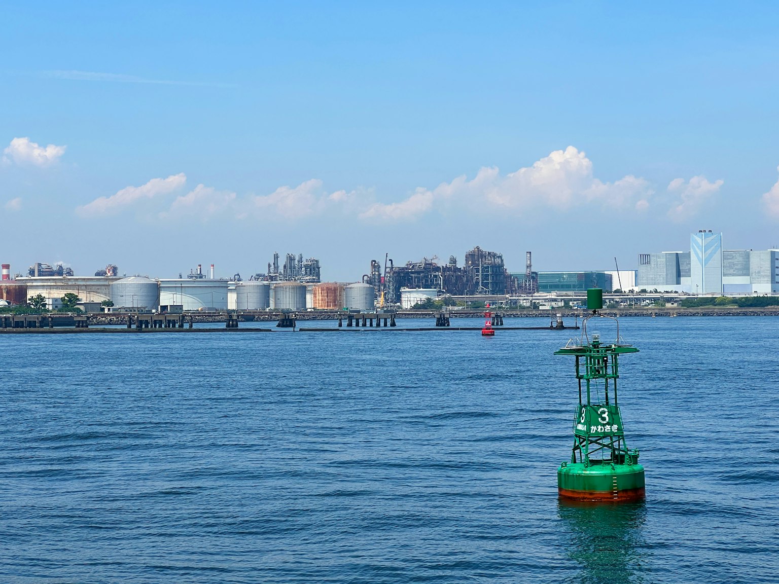 A green buoy floating on blue water with an industrial area in the background