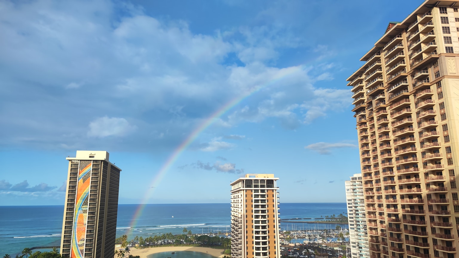 Rainbow arching between skyscrapers with a clear blue sky and ocean view