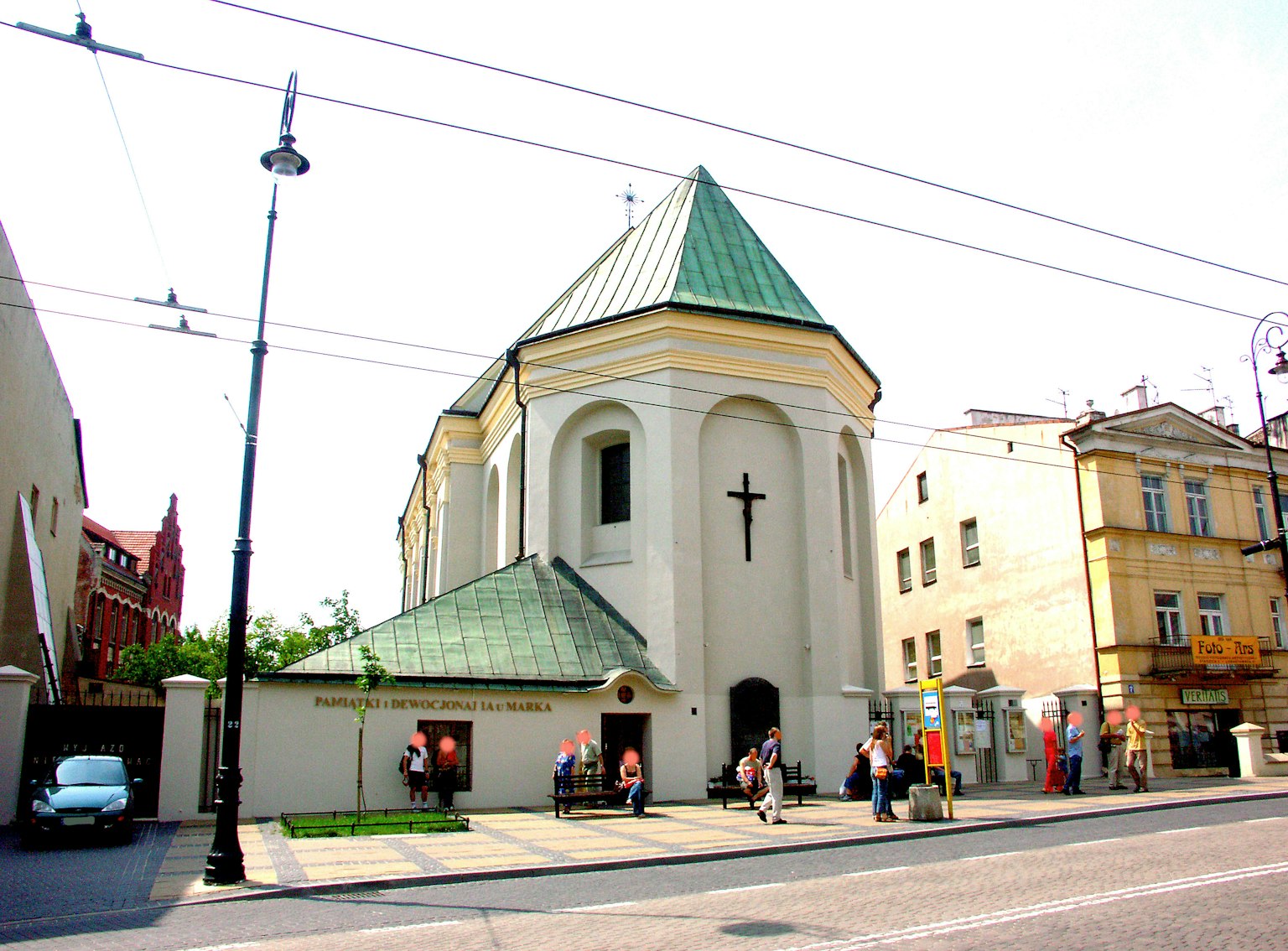 Esterno di una piccola chiesa con tetto verde e decorazione a croce persone e auto nelle vicinanze