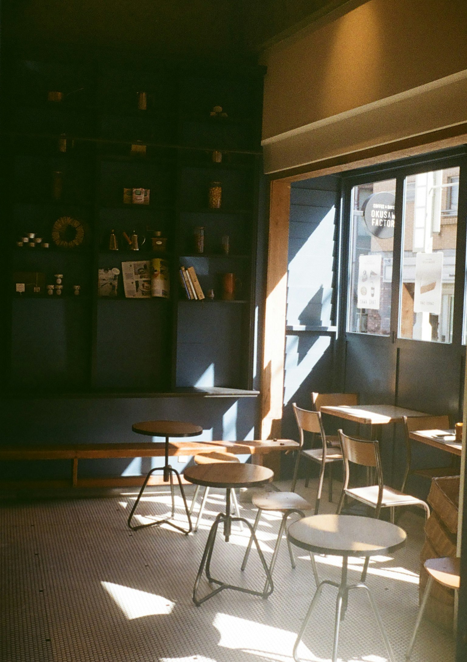 Interior de café bien iluminado con luz natural mesas y sillas de madera simples dispuestas estanterías en la pared con libros y objetos decorativos