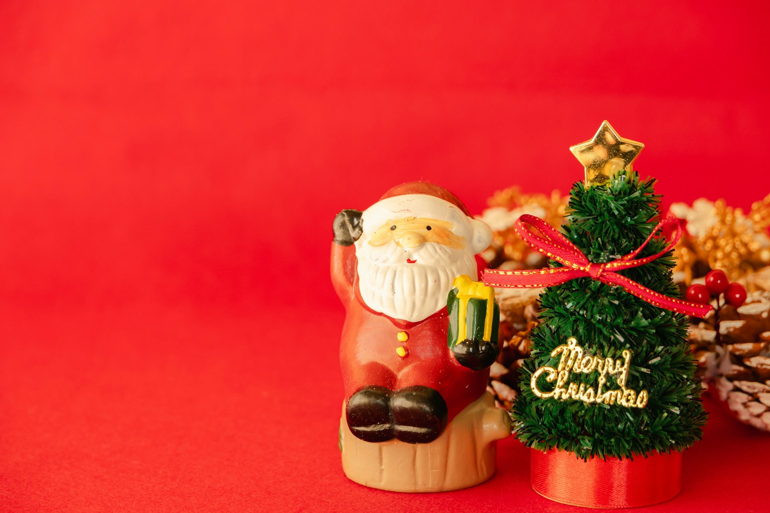Tượng ông già Noel ngồi bên cạnh cây thông Giáng sinh nhỏ trên nền đỏ