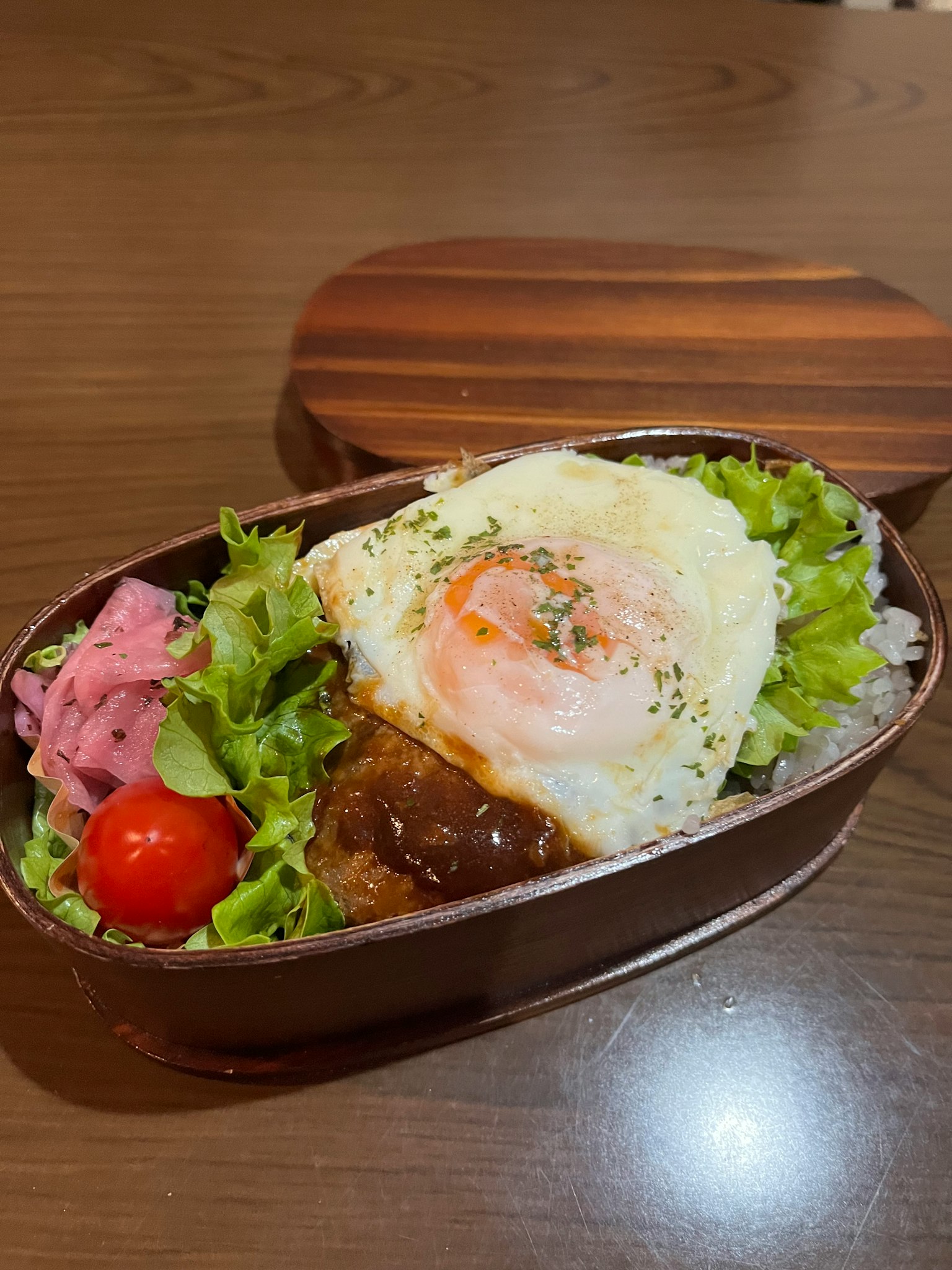 Caja bento con un huevo frito sobre arroz, ensalada, tomates cherry y hamburguesa