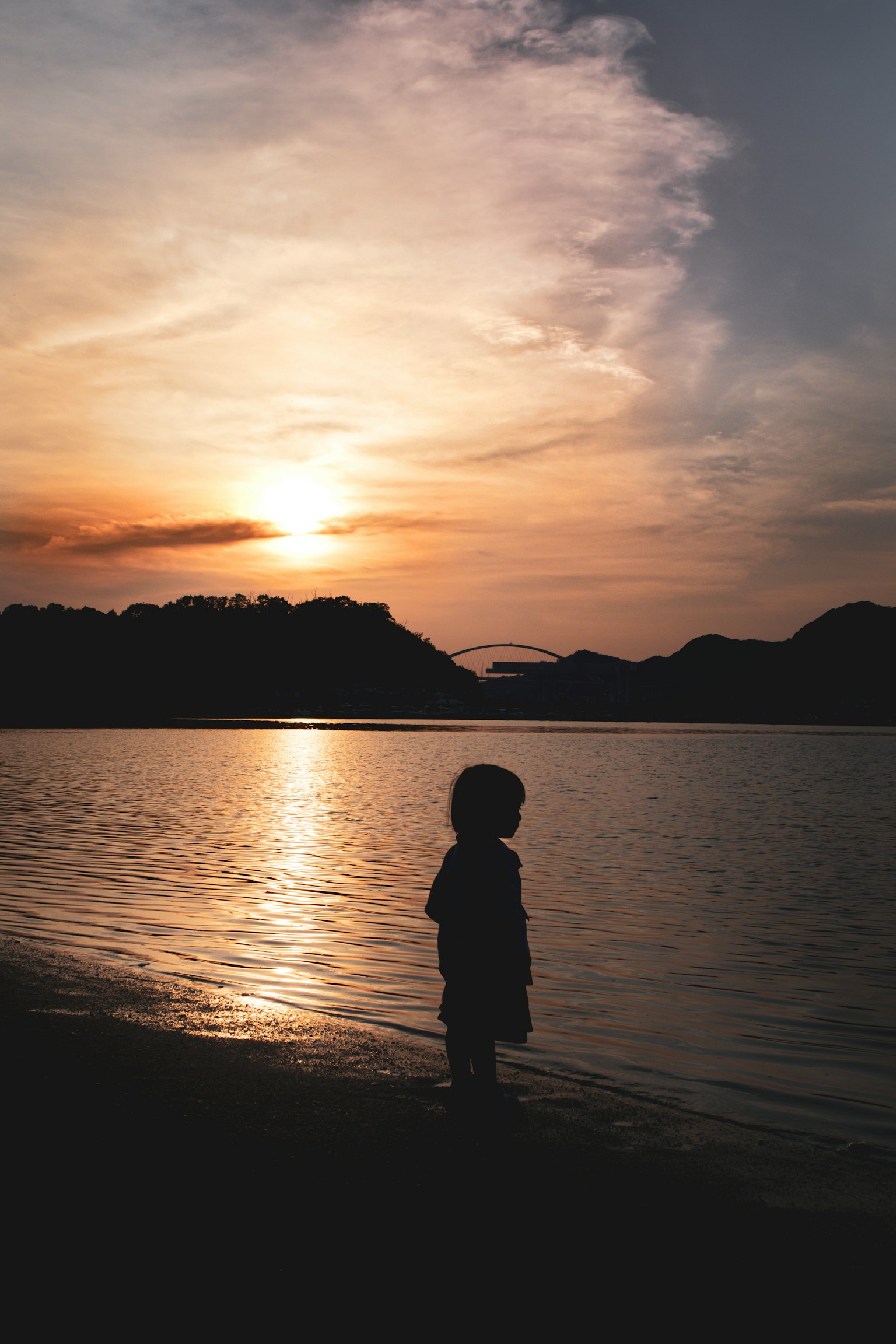 เงาของเด็กยืนอยู่ข้างน้ำในขณะที่พระอาทิตย์ตกดินมีภูเขาอยู่เบื้องหลัง