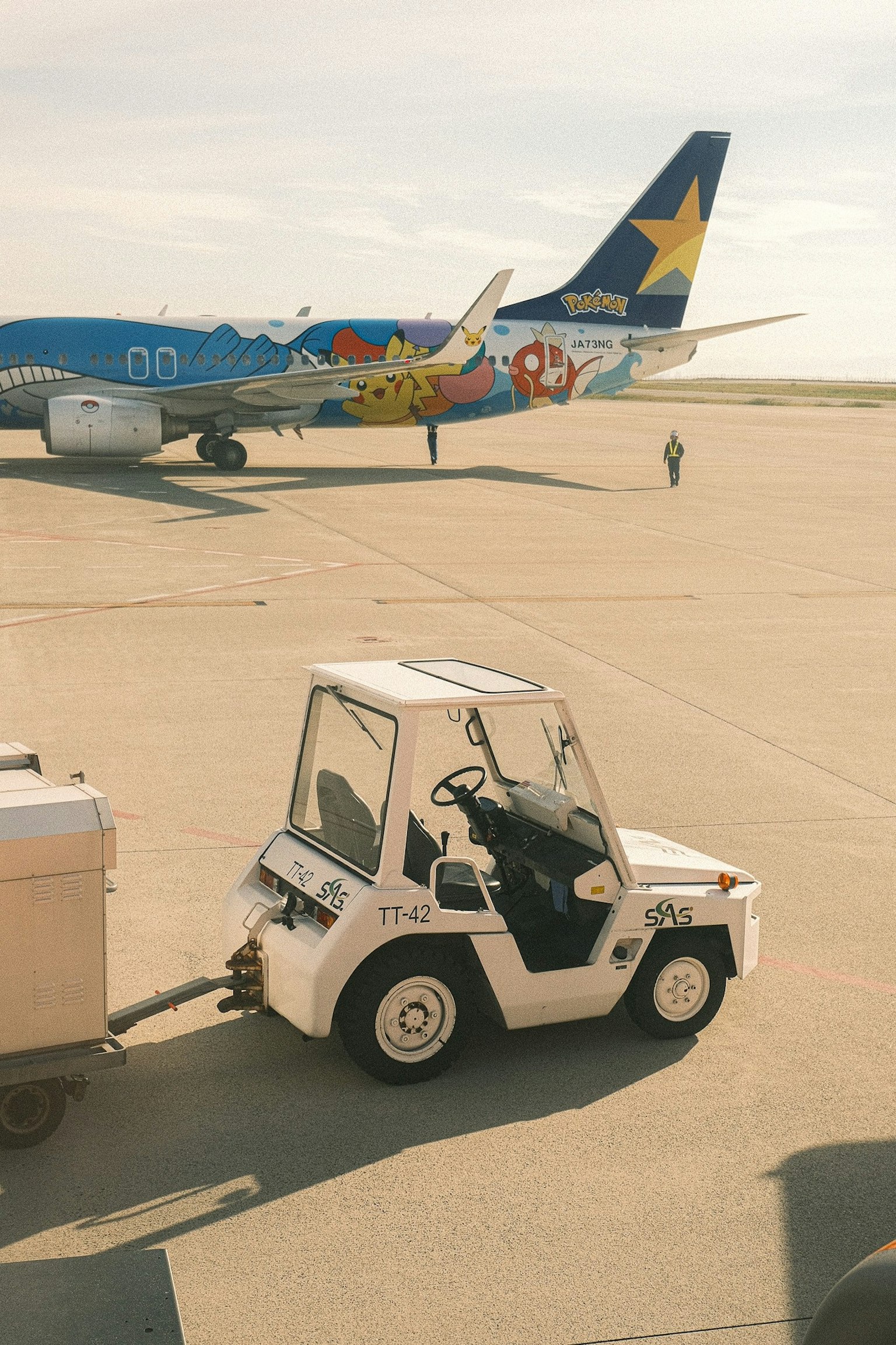 Weißes Abschleppfahrzeug auf dem Flughafen-Rollfeld mit buntem Flugzeug im Hintergrund
