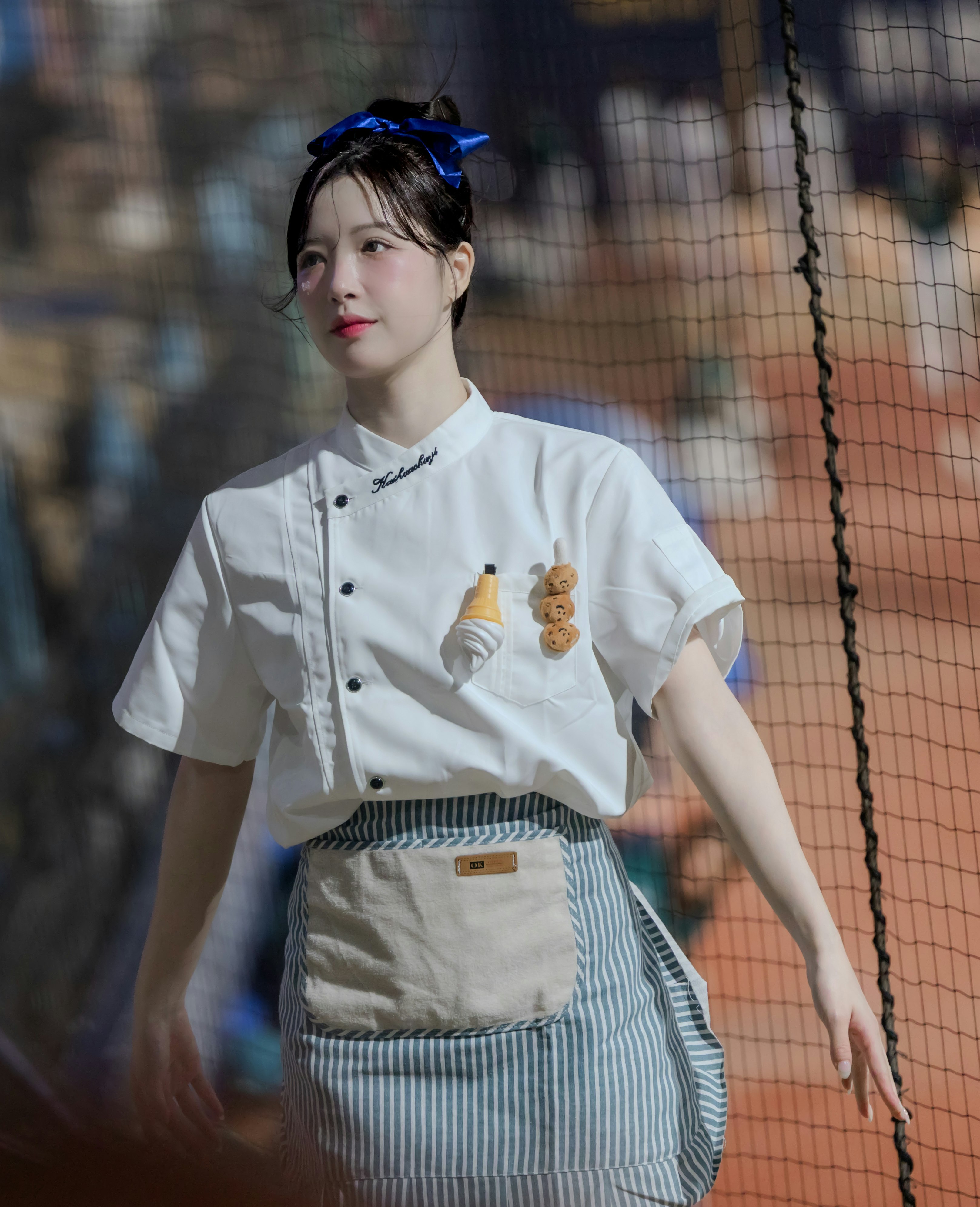 白い制服風の衣装を着た女性がフェンス前で微笑む