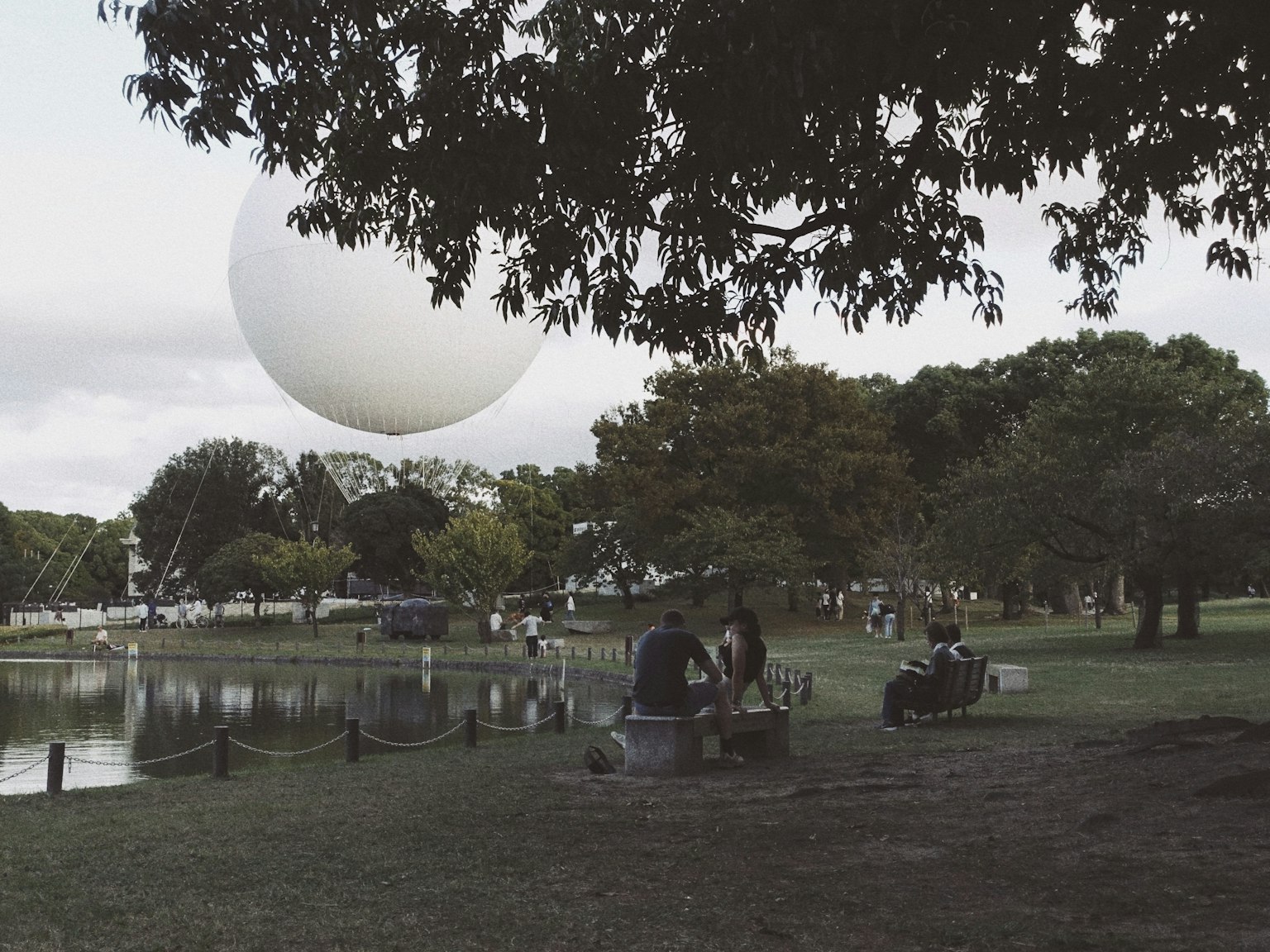 Personas relajándose cerca de un lago en un parque con una gran esfera blanca flotando en el cielo
