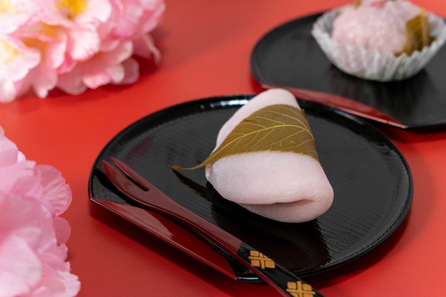 Dolci giapponesi su un piatto nero con bacchette circondati da fiori rosa e uno sfondo rosso