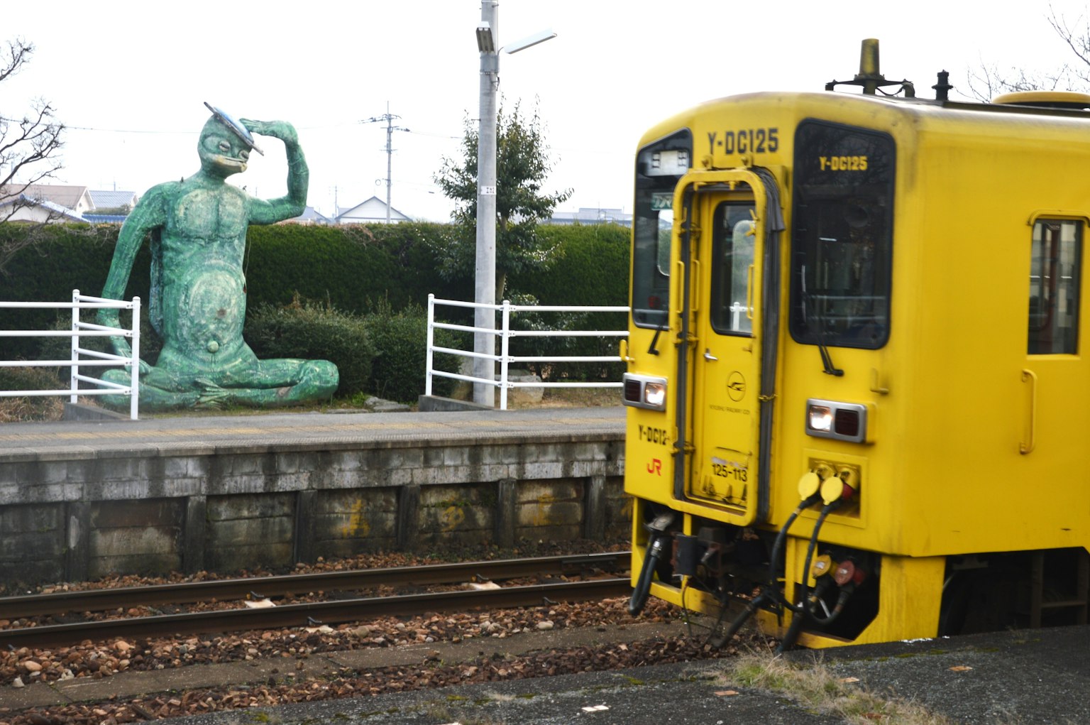 Gelber Zug an einer Station mit einer grünen Skulptur