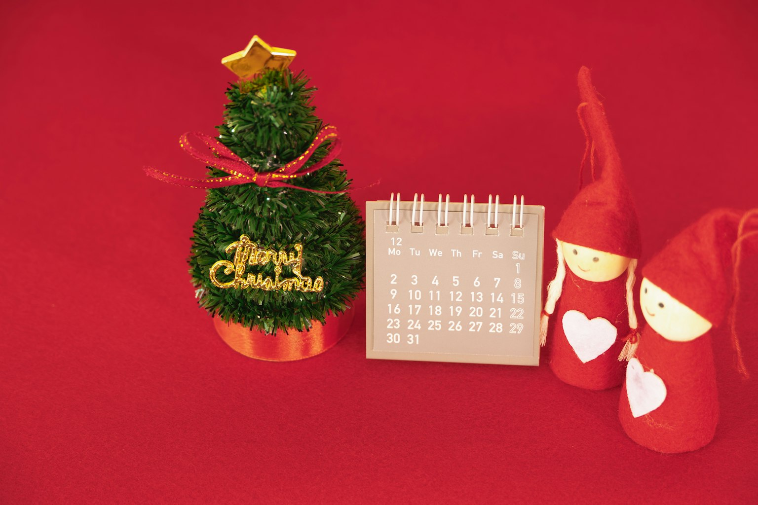 Un piccolo albero di Natale con una stella e un calendario accanto a due bambole festive su uno sfondo rosso