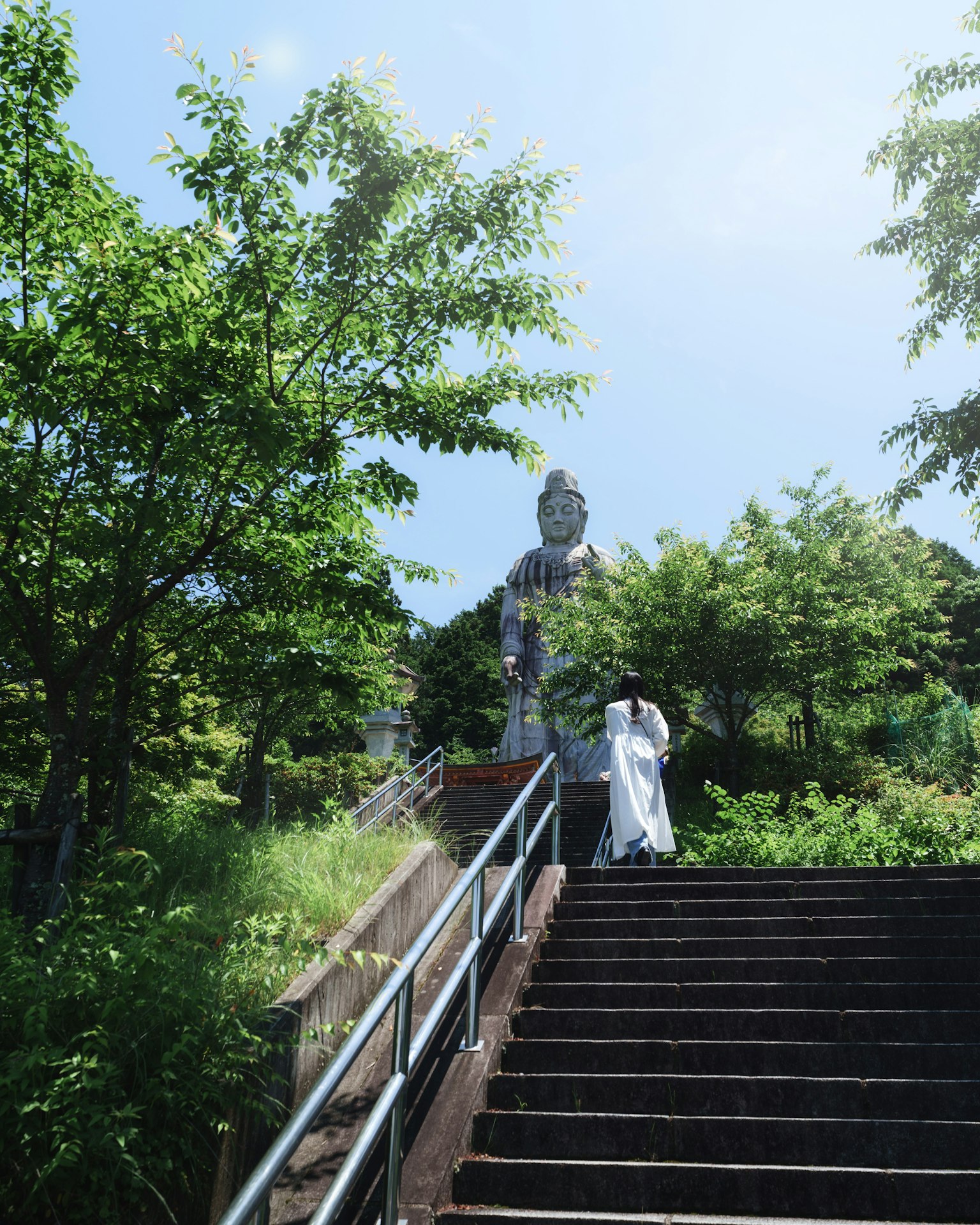 Una persona con ropa blanca subiendo escaleras de piedra rodeadas de vegetación con una gran estatua de Buda al fondo