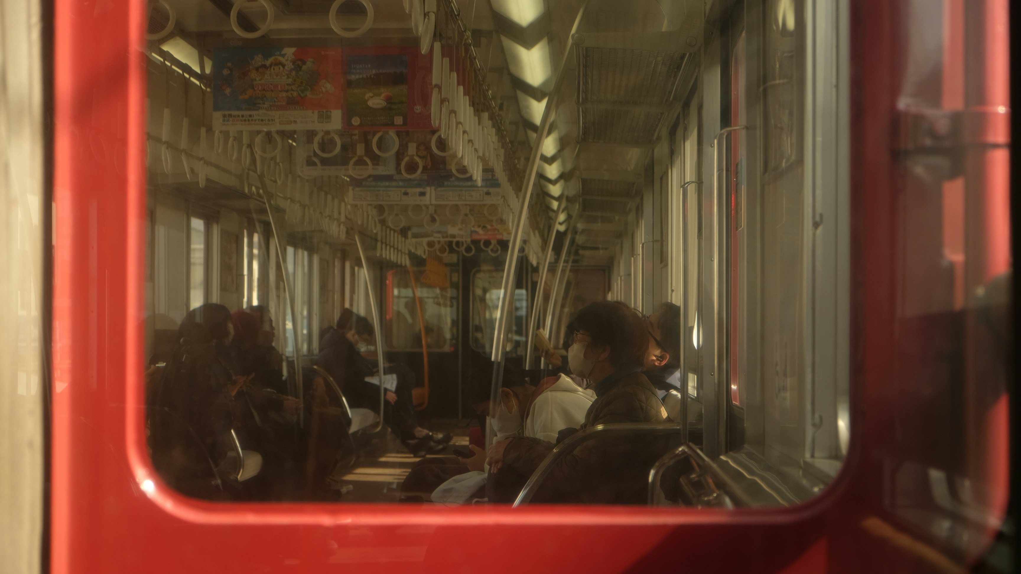 赤い車両の窓越しに人が座る地下鉄の車内