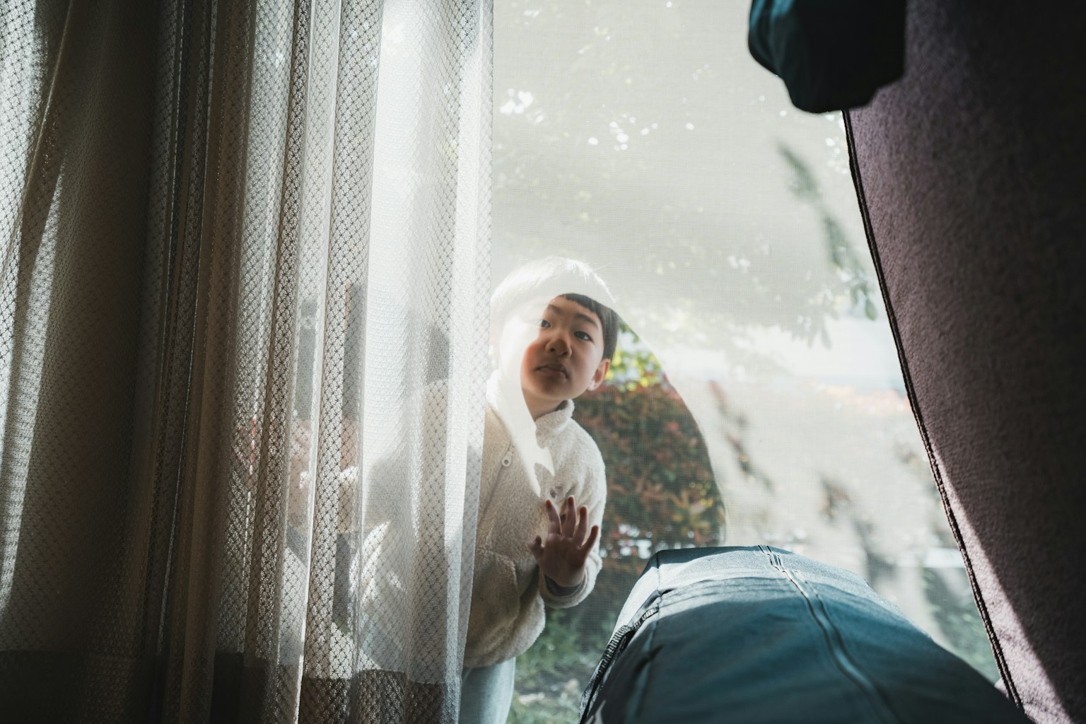 A surprised child seen through sheer curtains with greenery outside
