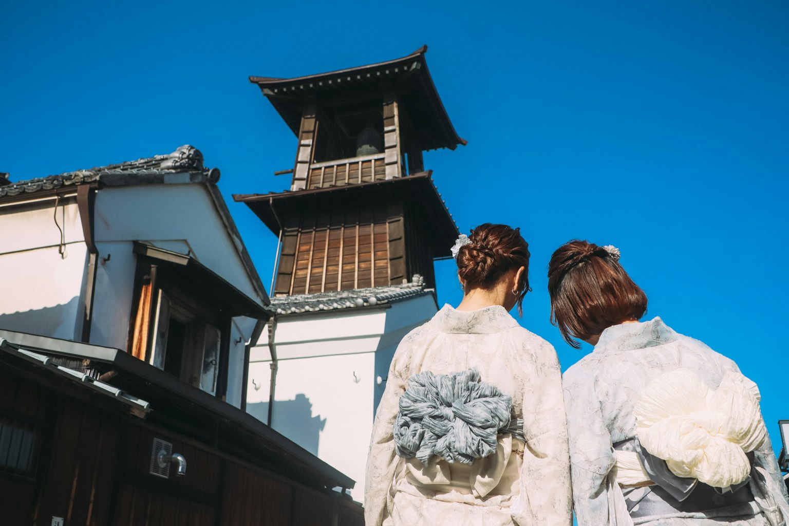 Two women in traditional attire looking back at an old tower under a blue sky