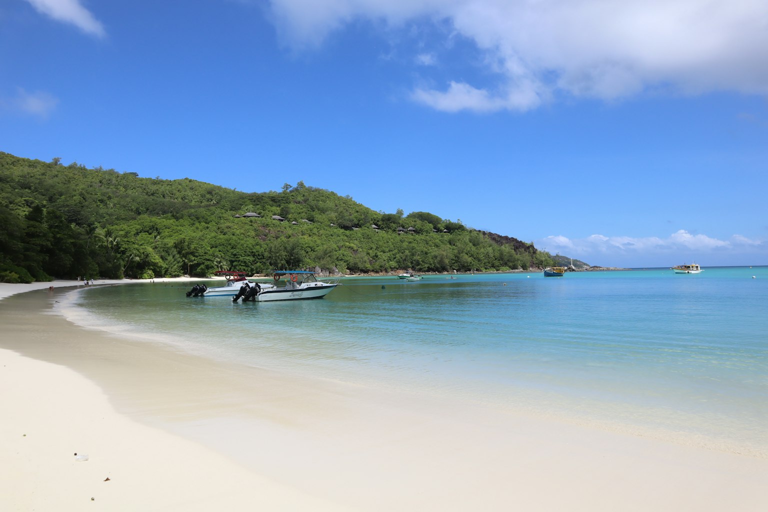 风景如画的海滩景观 清澈的蓝色海水和白色沙滩 附近停靠的船只 背景是郁郁葱葱的绿色山丘