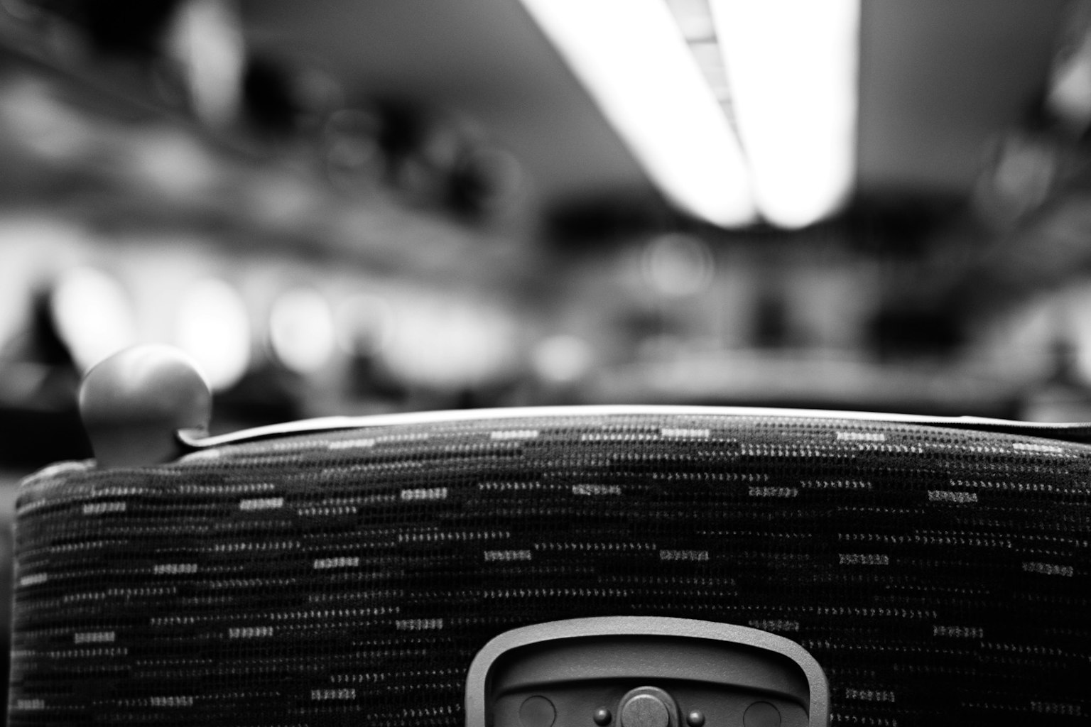 モノクロの電車の座席の背もたれが前景にあり、背景にはぼやけた乗客と照明が見える