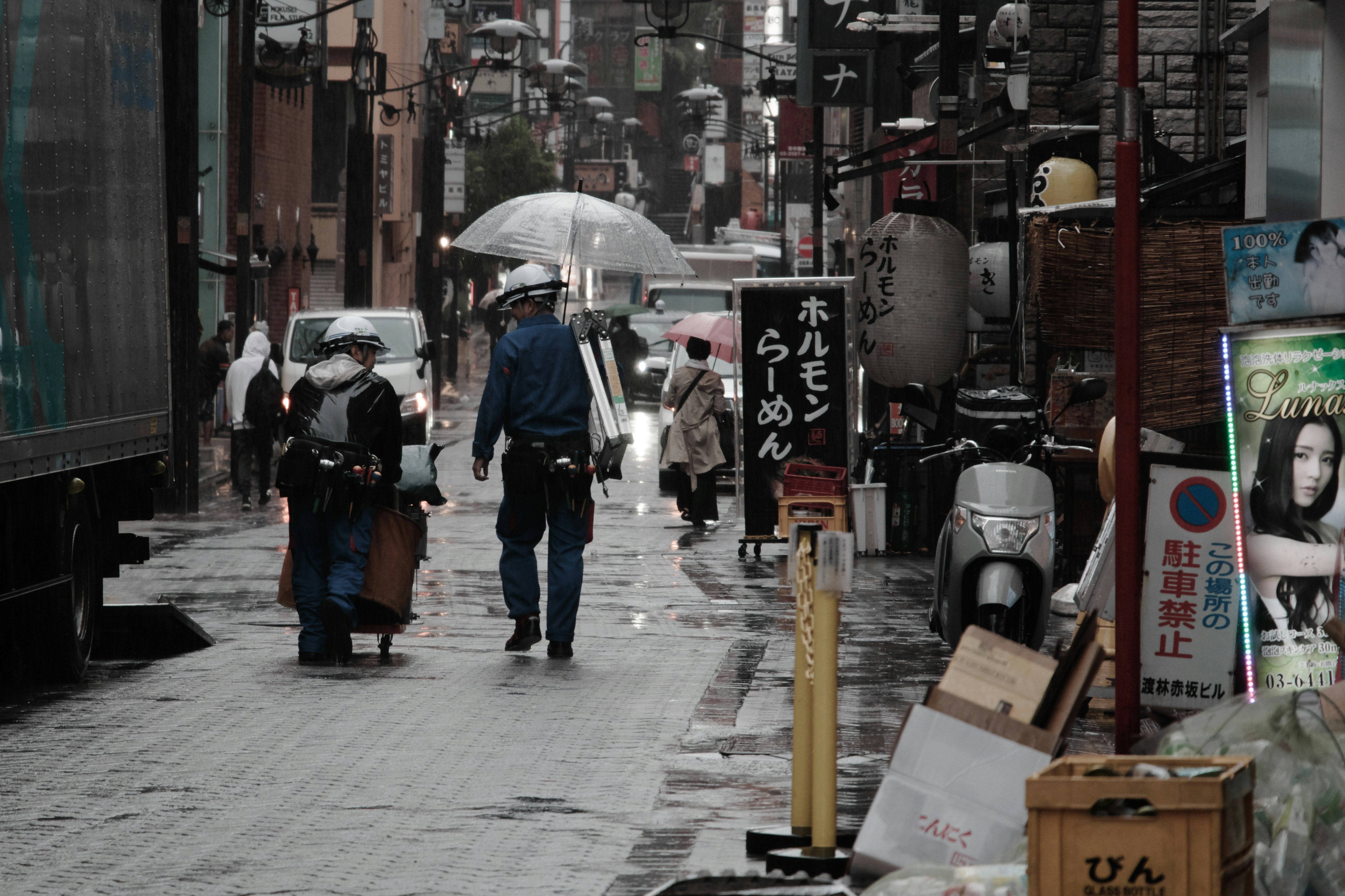 雨の路地で傘をさす人と軒先の看板が並ぶ商店街