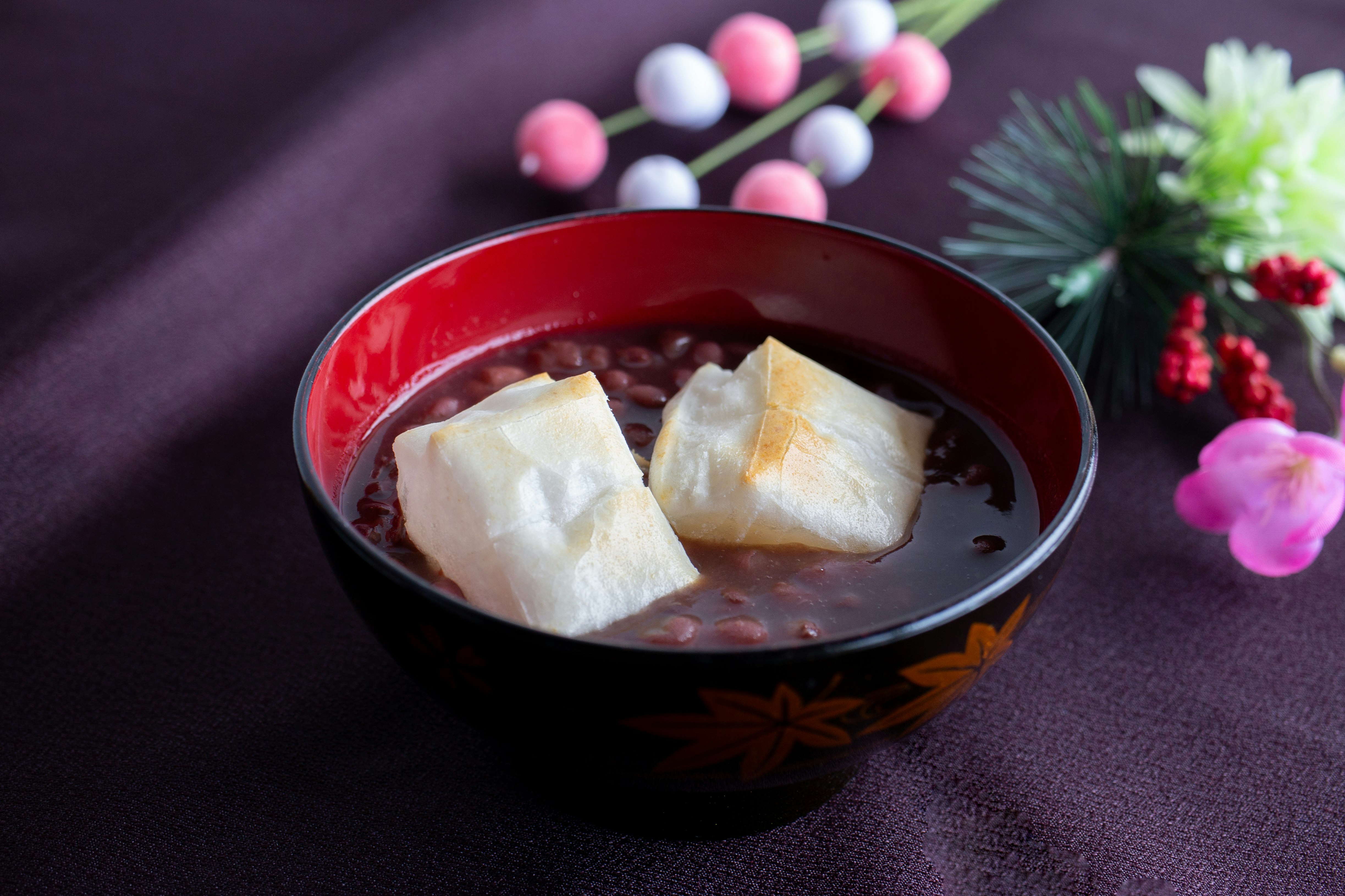 Sopa dulce con mochi en cubos en un cuenco rojo, decorada con pino y flores