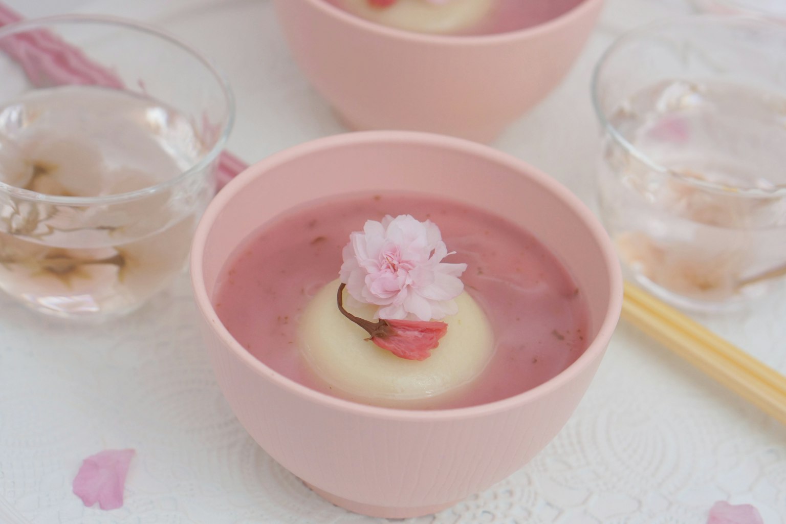 Una hermosa disposición de mesa con un postre rosa con una flor de cerezo flotante y vasos transparentes al lado