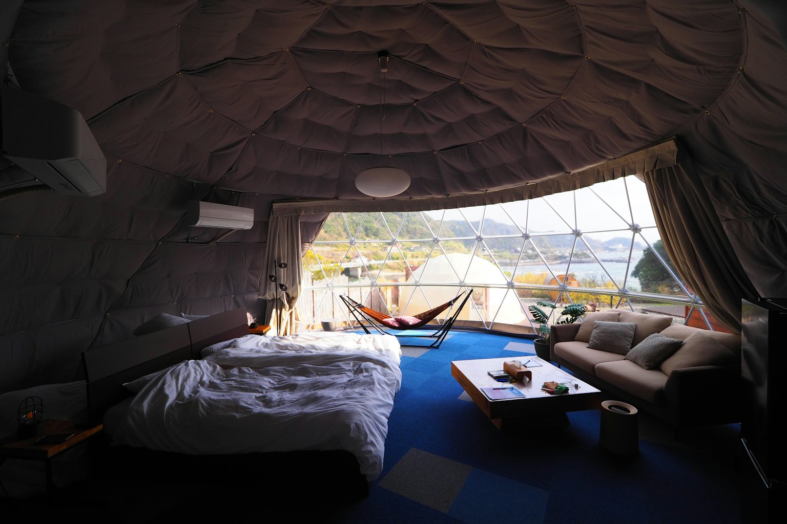 Interior of a dome-shaped accommodation featuring a bed and sofa with a scenic view outside