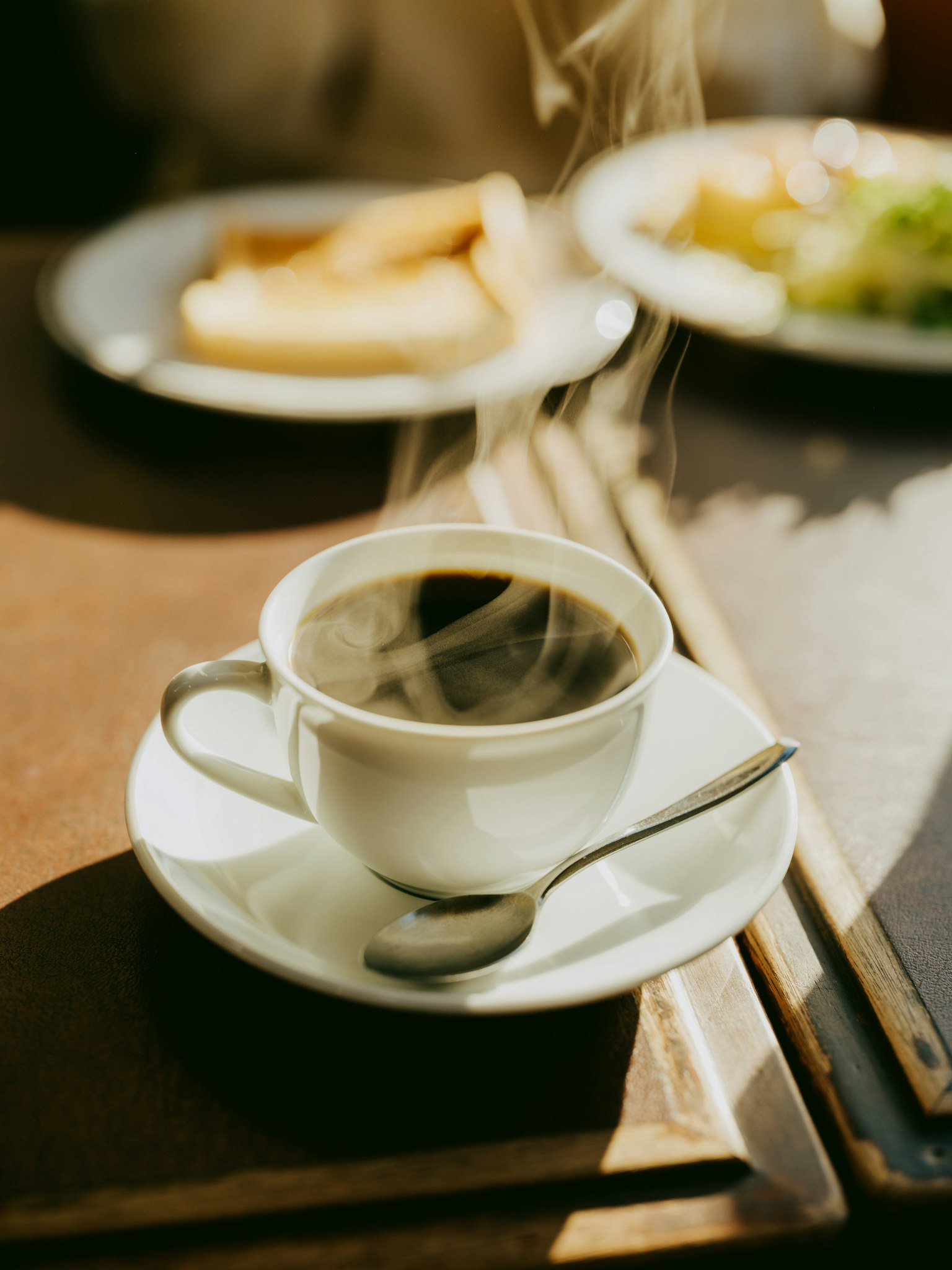 Một cốc cà phê nóng trong cốc trắng với thìa bên cạnh đĩa thức ăn