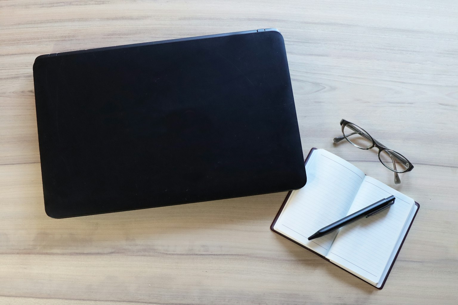 Laptop đen bên cạnh sổ tay và bút với kính trên bàn gỗ