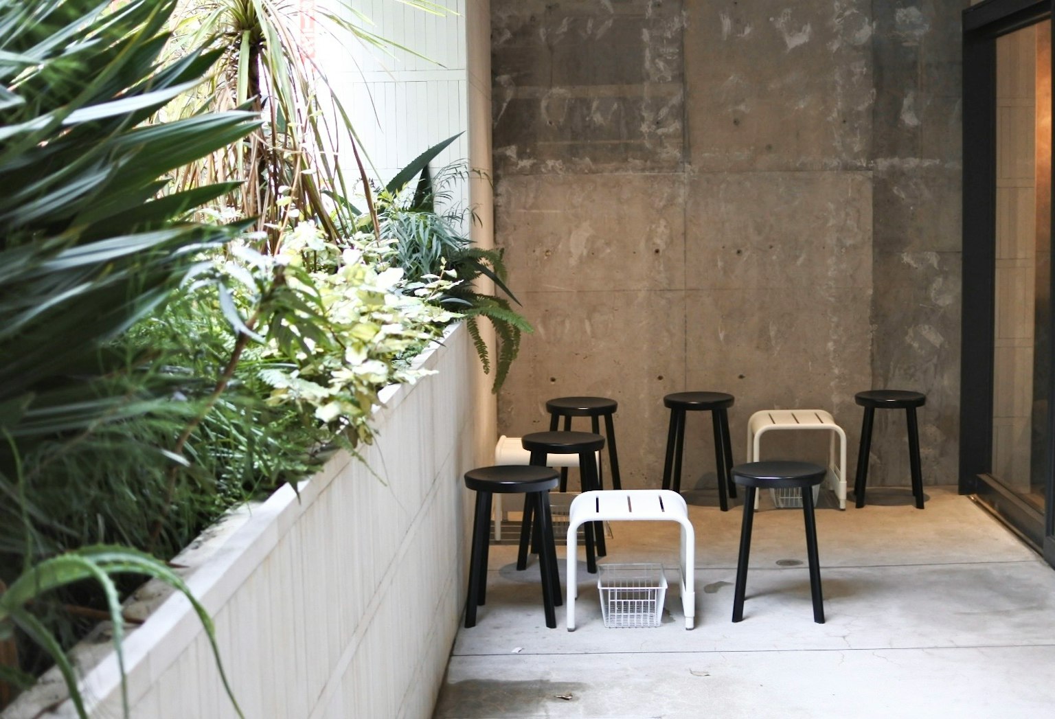 Una terraza con una pared de concreto que presenta taburetes negros y blancos rodeados de vegetación