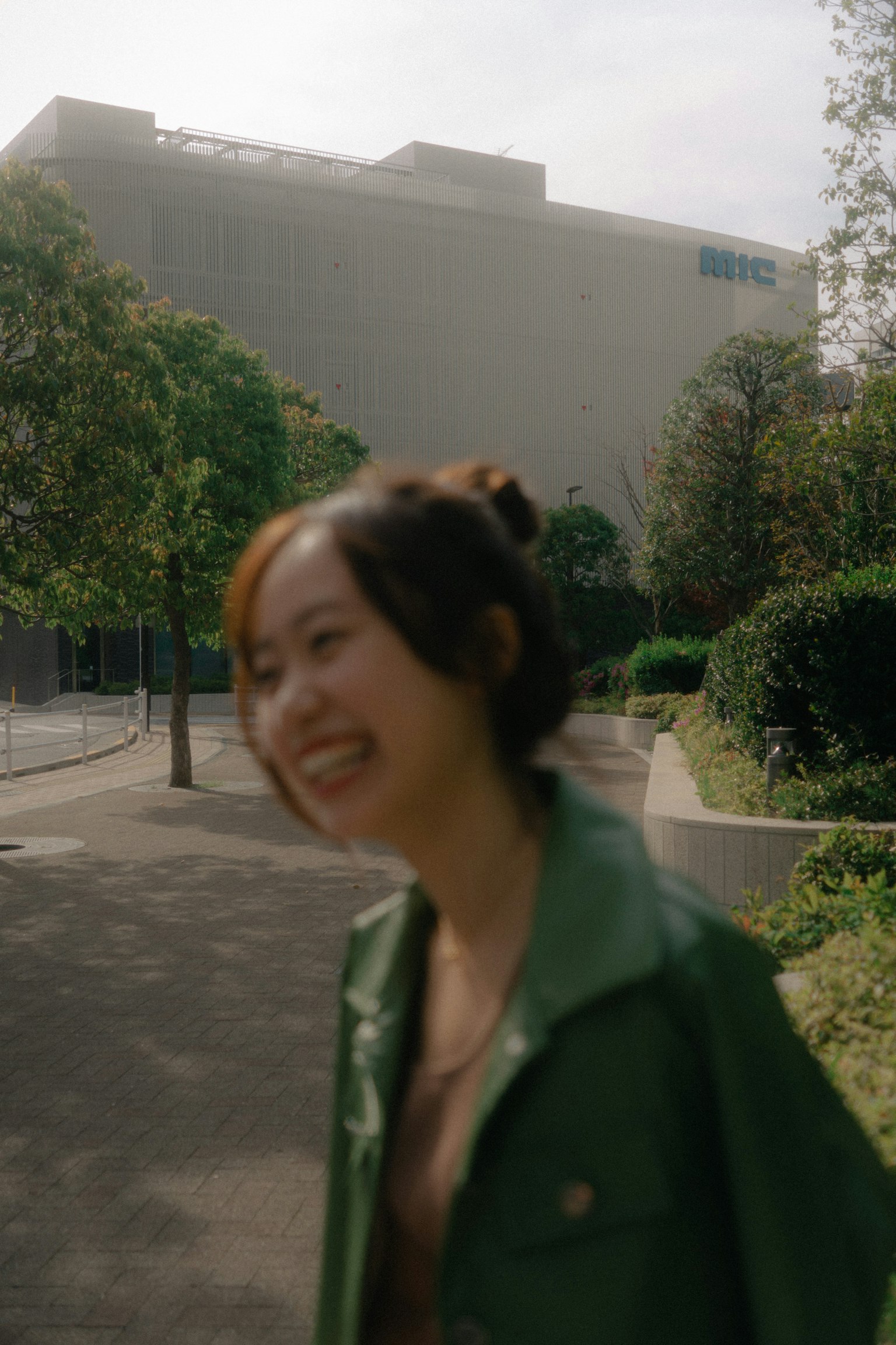 Una mujer sonriente con chaqueta verde caminando rodeada de árboles y un edificio al fondo