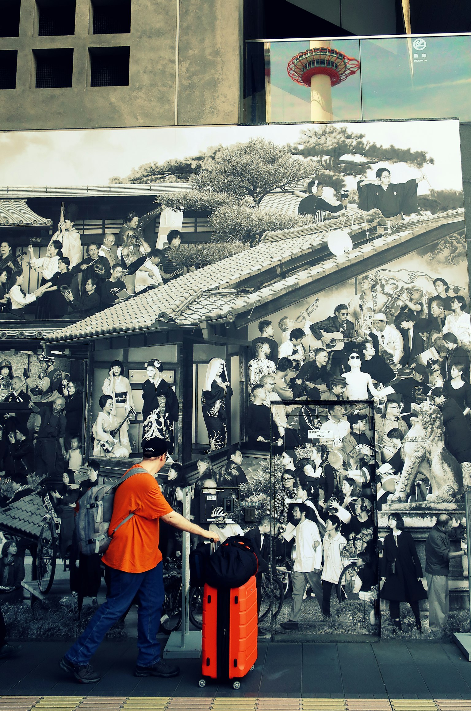 Du khách mặc áo sơ mi cam đẩy vali đỏ trước bức tranh tường đen trắng mô tả cảnh đông đúc