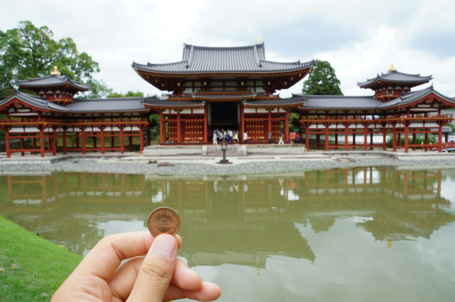 Mano sosteniendo una moneda frente a un edificio japonés tradicional junto a un estanque