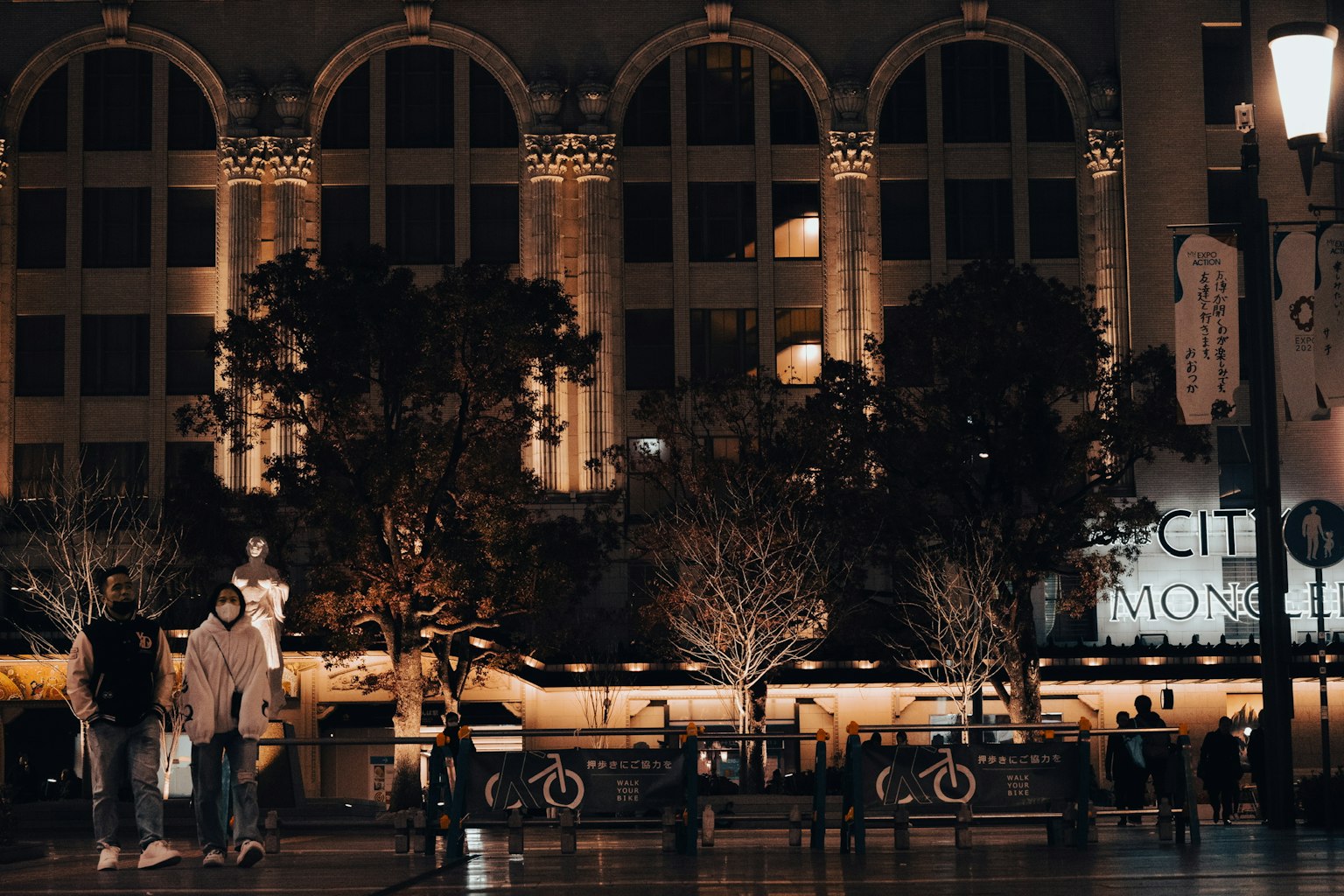 People gathering in a nighttime urban setting featuring large arched buildings and lined trees