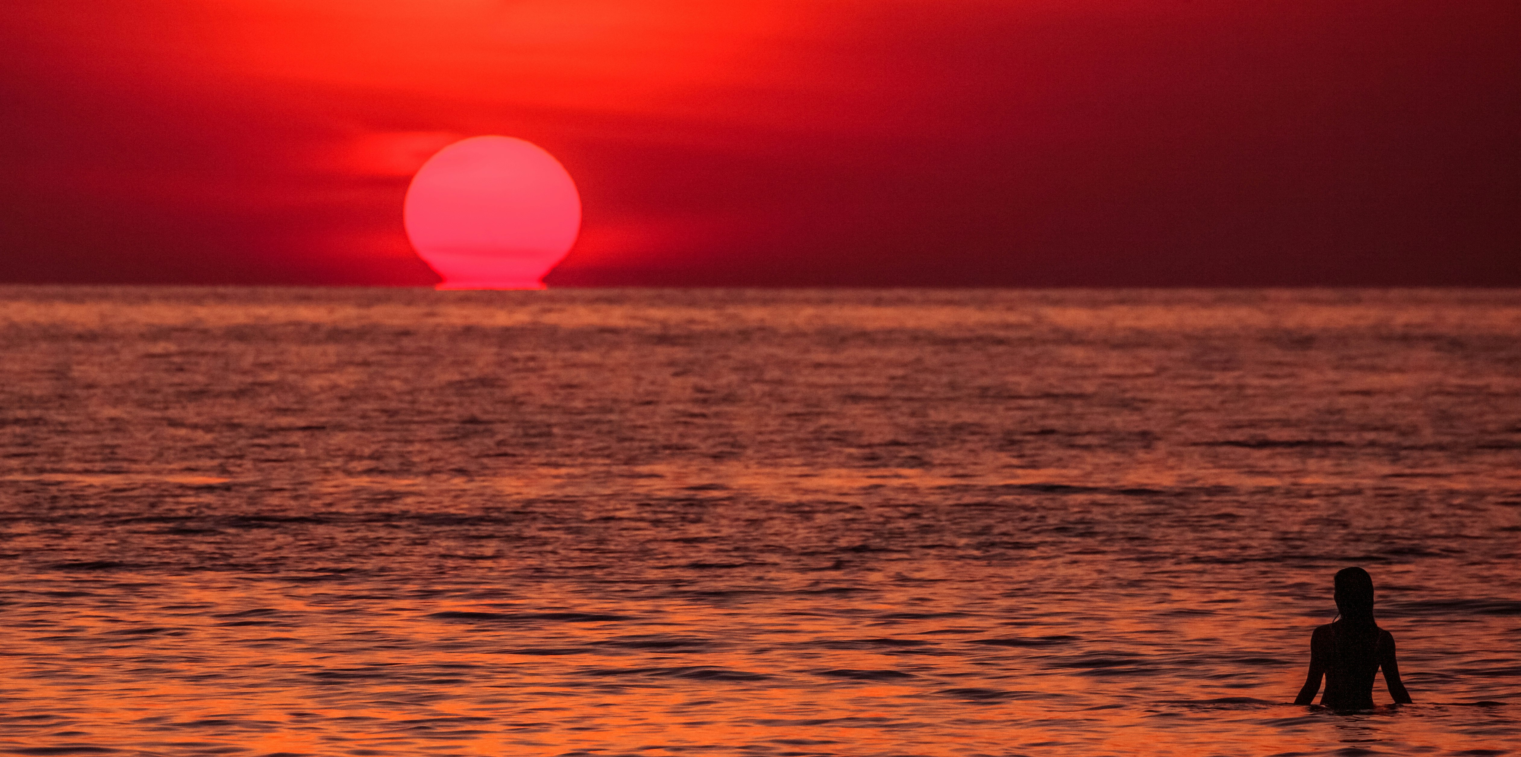 夕焼けの海辺で一人の人物が波打ち際に立つ