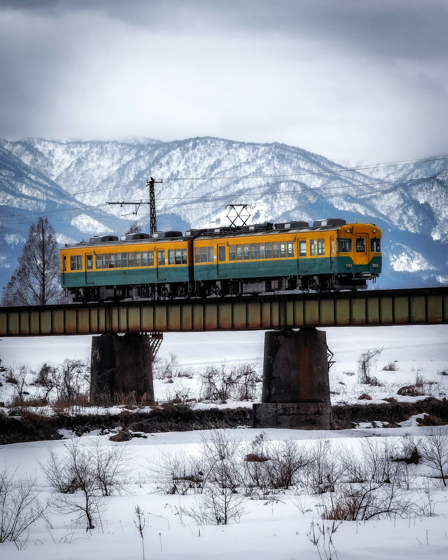 Treno giallo che attraversa un ponte in un paesaggio innevato con montagne