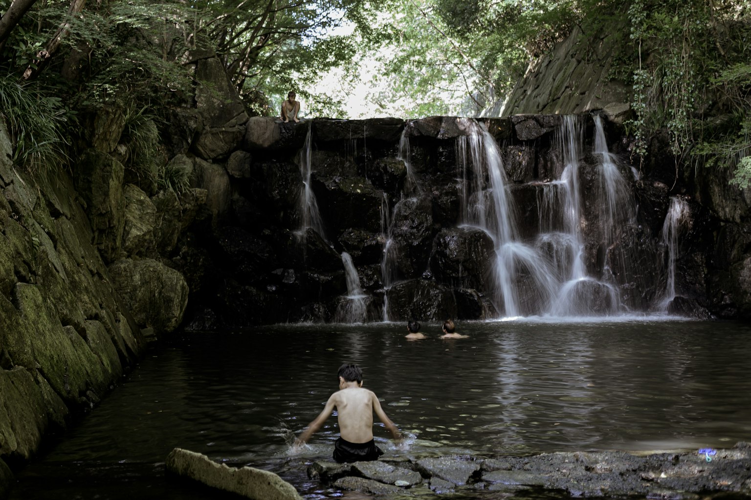 ชายคนหนึ่งนั่งอยู่ใกล้น้ำตกในสภาพแวดล้อมทางธรรมชาติที่เงียบสงบล้อมรอบด้วยพืชพรรณเขียวขจีและหิน