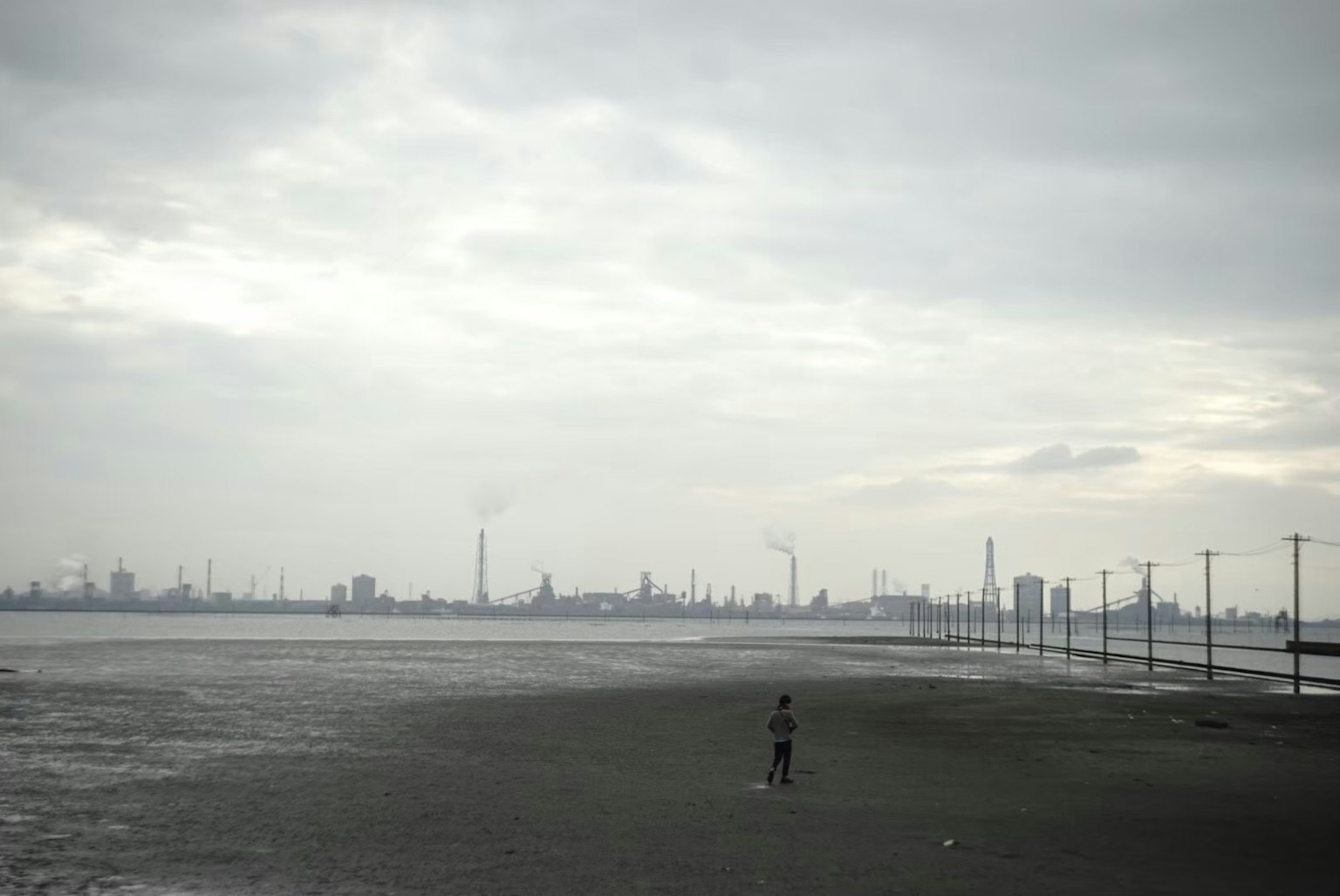 Sosok sendirian di pantai dengan garis langit industri di bawah langit mendung
