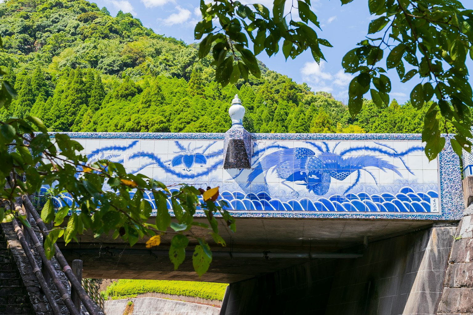 Jembatan dengan desain gelombang biru dan tiang dekoratif putih dikelilingi oleh pegunungan hijau