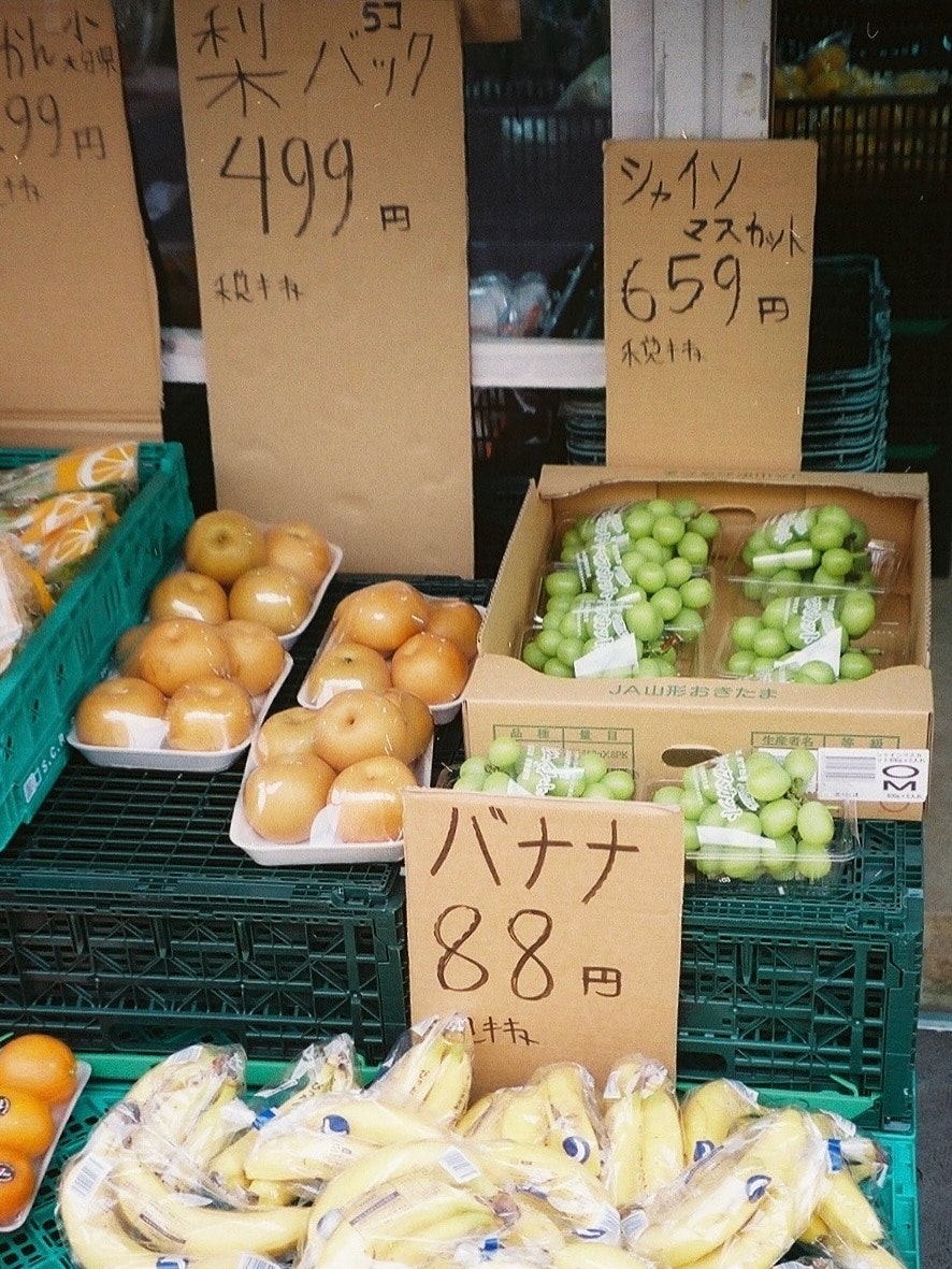 青いプラスチックかごに並ぶ果物と値札のある店先