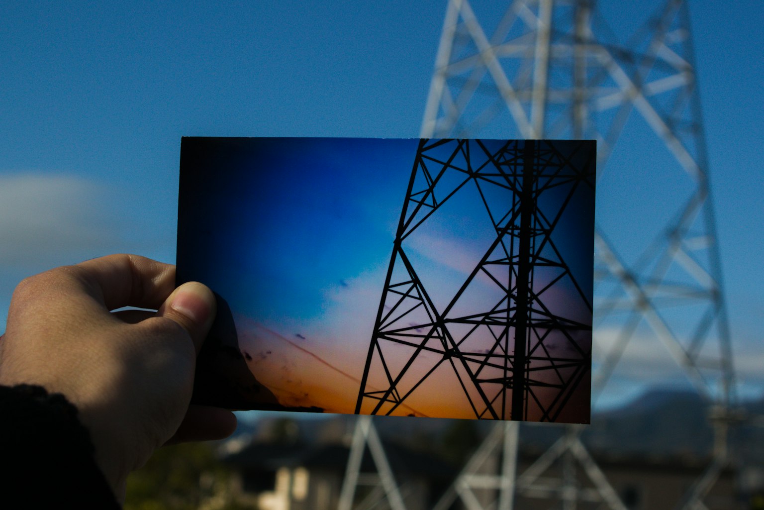 A hand holding a landscape photo with a power tower in the background blue sky and orange gradient