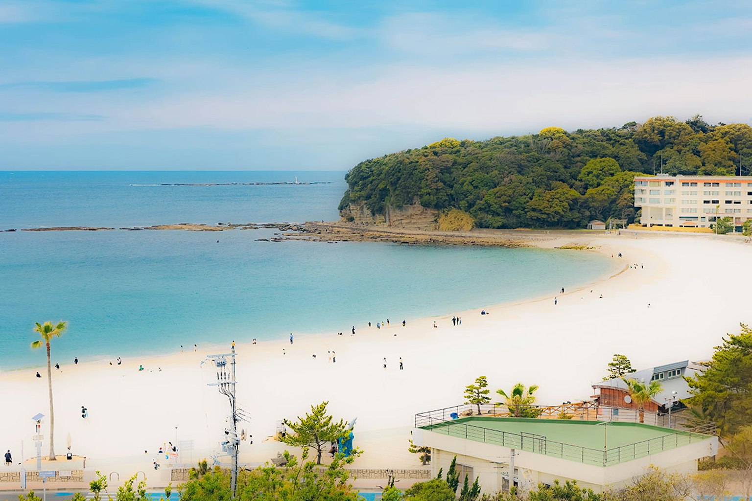 Malersicher Blick auf einen ruhigen Strand mit blauem Meer Menschen verstreut am Sandstrand grüne Hügel und Gebäude im Hintergrund