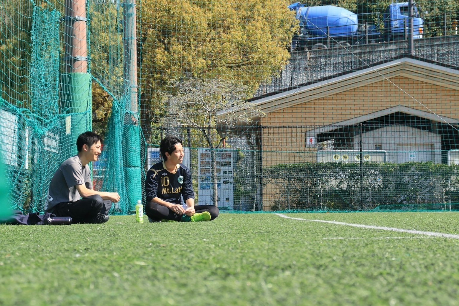 Dua pria duduk di atas rumput sintetis di lapangan sepak bola