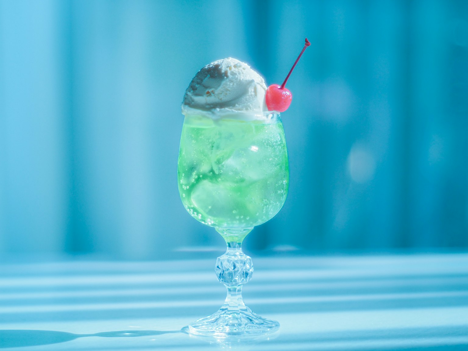 A green cocktail in a blue glass topped with ice cream and a cherry