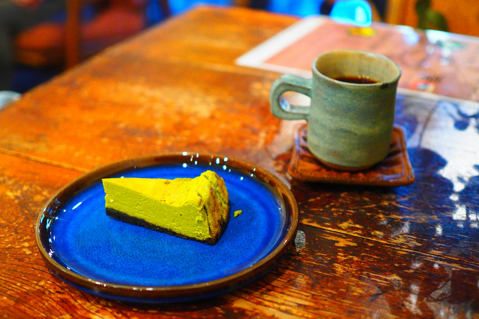 Una fetta di torta gialla su un piatto blu accanto a una tazza grigia di caffè su un tavolo di legno