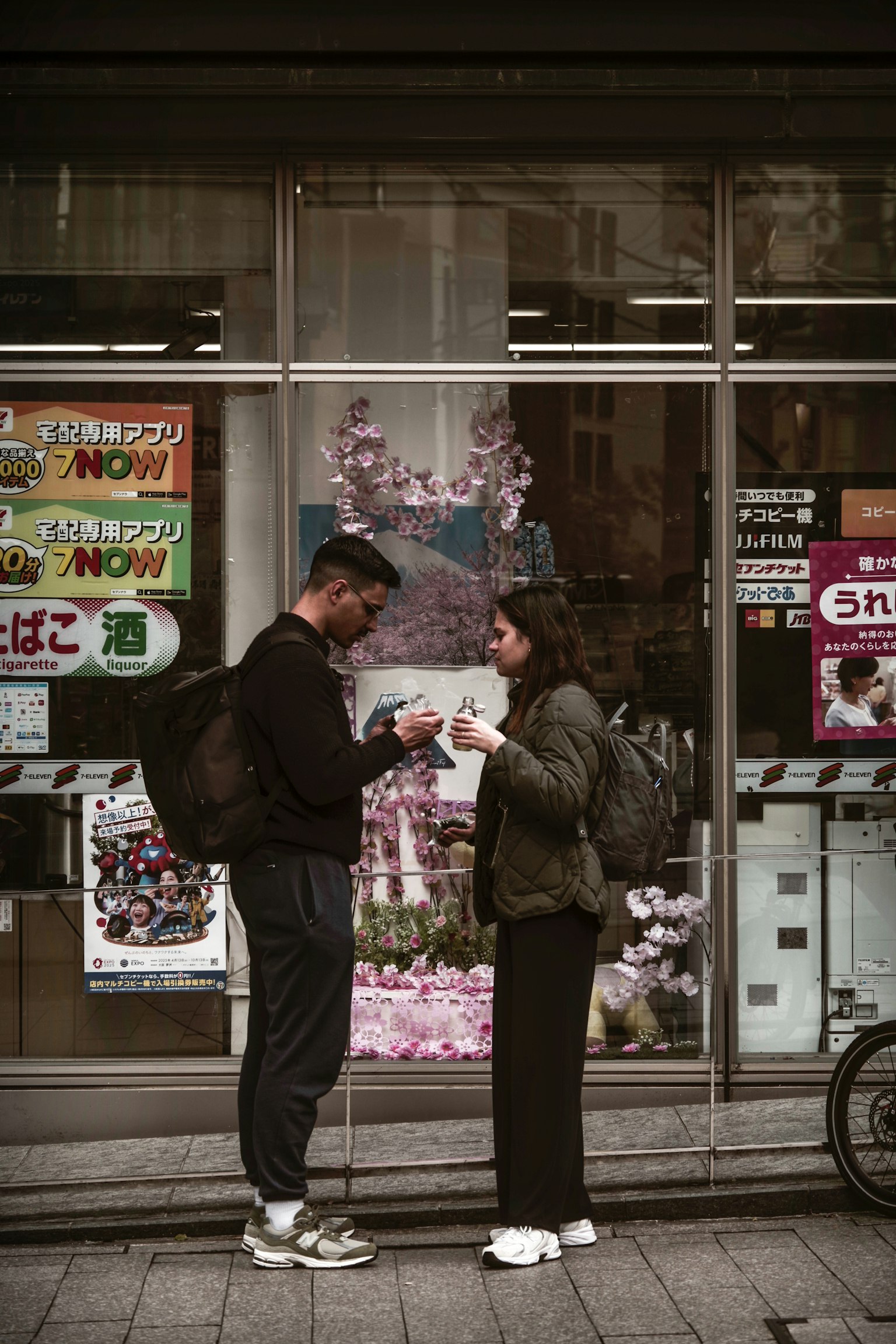 Một cặp đôi ăn mặc thoải mái đang trao đổi đồ uống trước cửa hàng có trang trí hoa anh đào và nhiều áp phích khác nhau