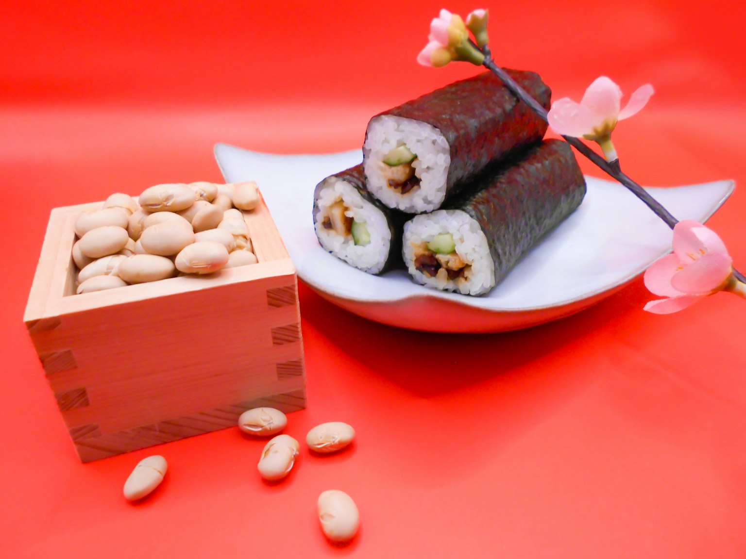 Sushi gulung di atas piring putih dengan kotak kayu berisi kacang dan bunga merah jambu di latar belakang merah